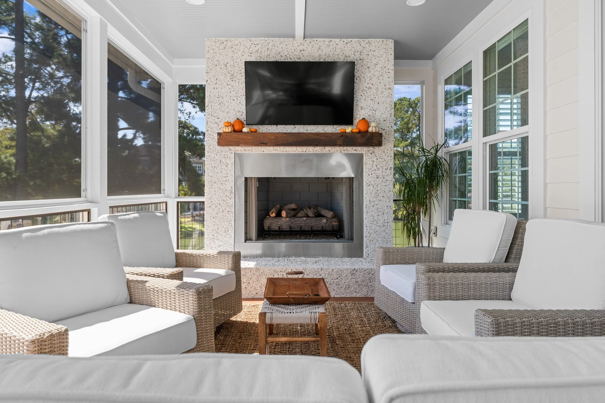 A cozy sunlit living room with a stone fireplace, a flat-screen TV above it, and white seating around a small wooden table, surrounded by large windows showing a green outdoor area.