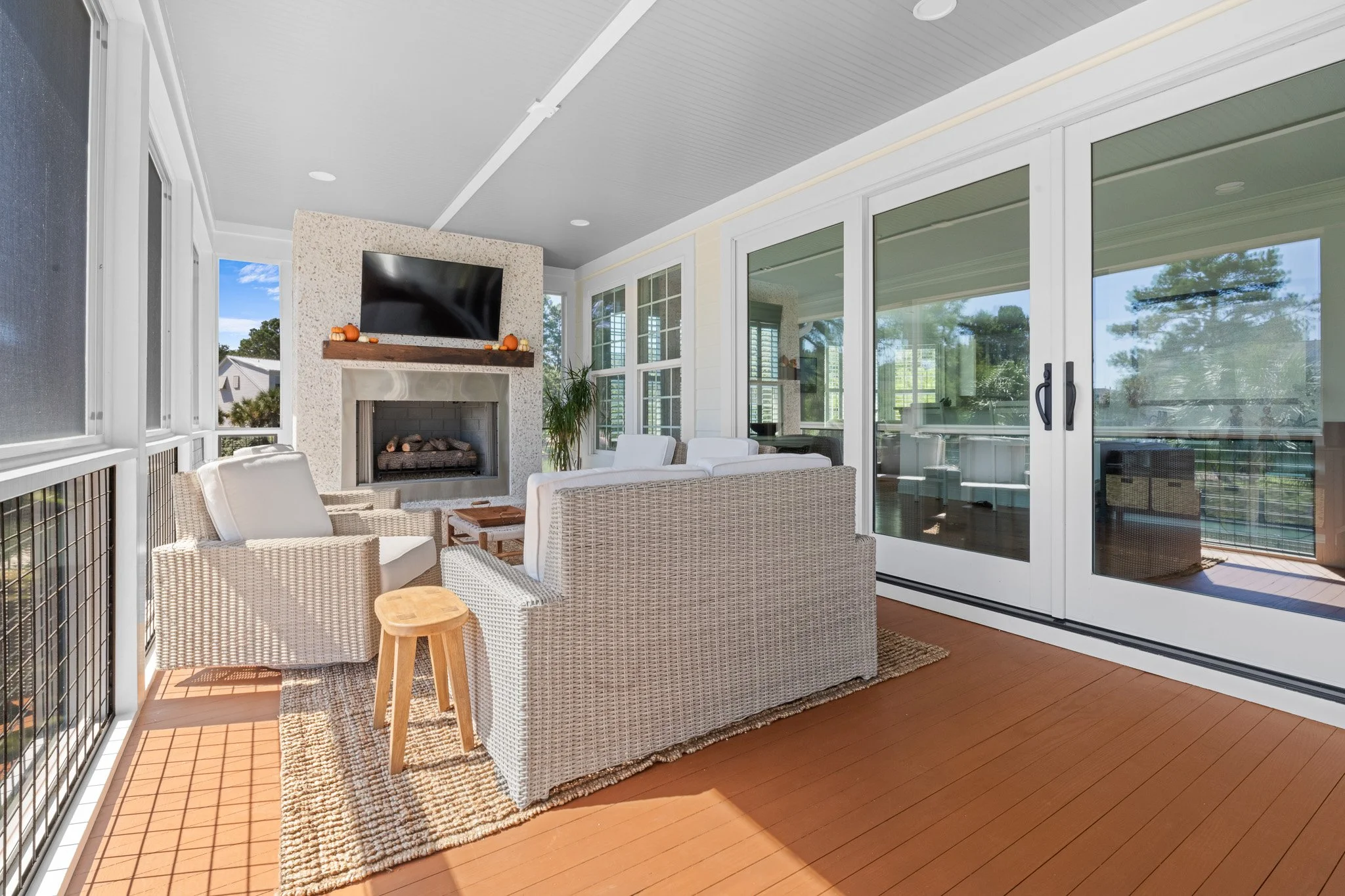 Sunlit enclosed porch with wicker furniture, a television above the fireplace, glass sliding doors, and a view of trees outside.