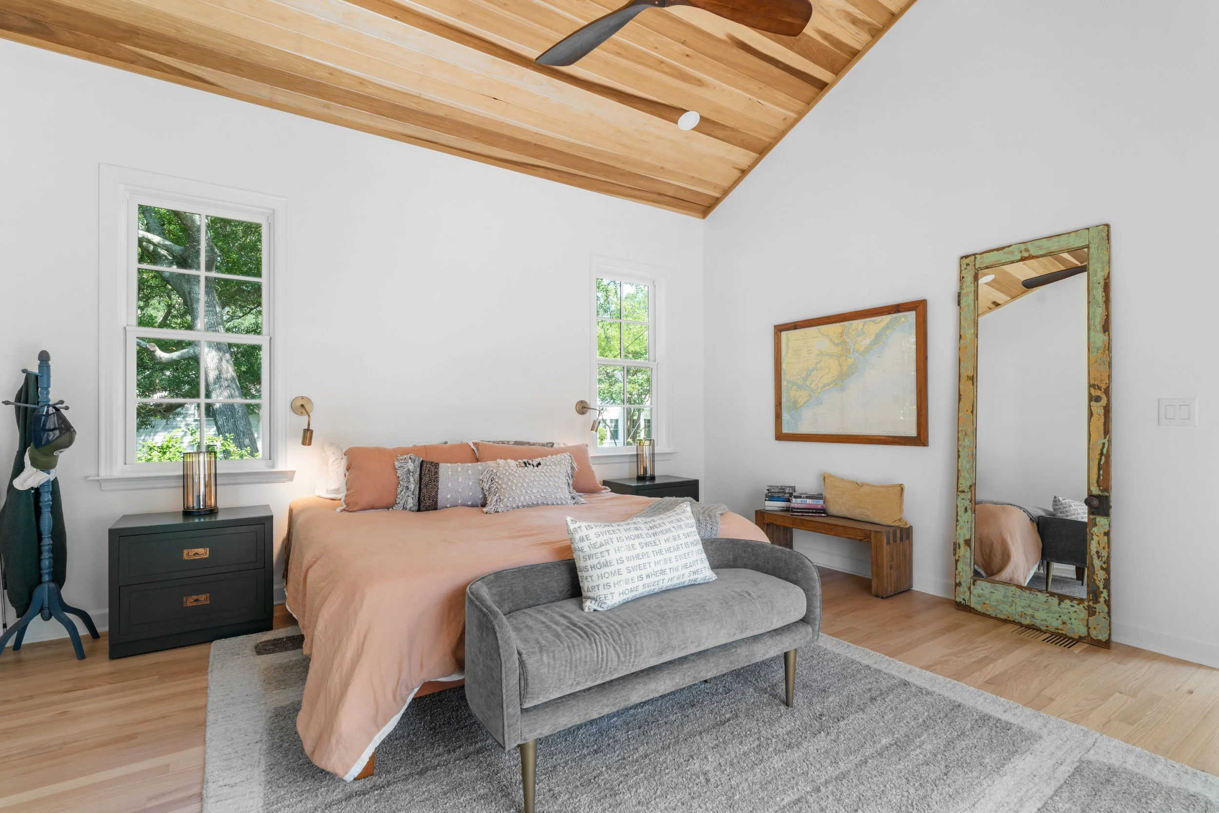 A bedroom with a wooden ceiling, two windows, a bed with peach bedding and various pillows, a gray bench at the foot of the bed, two black nightstands with lamps, a large mirror leaning against the wall, a wooden bench with cushions, a framed map on 