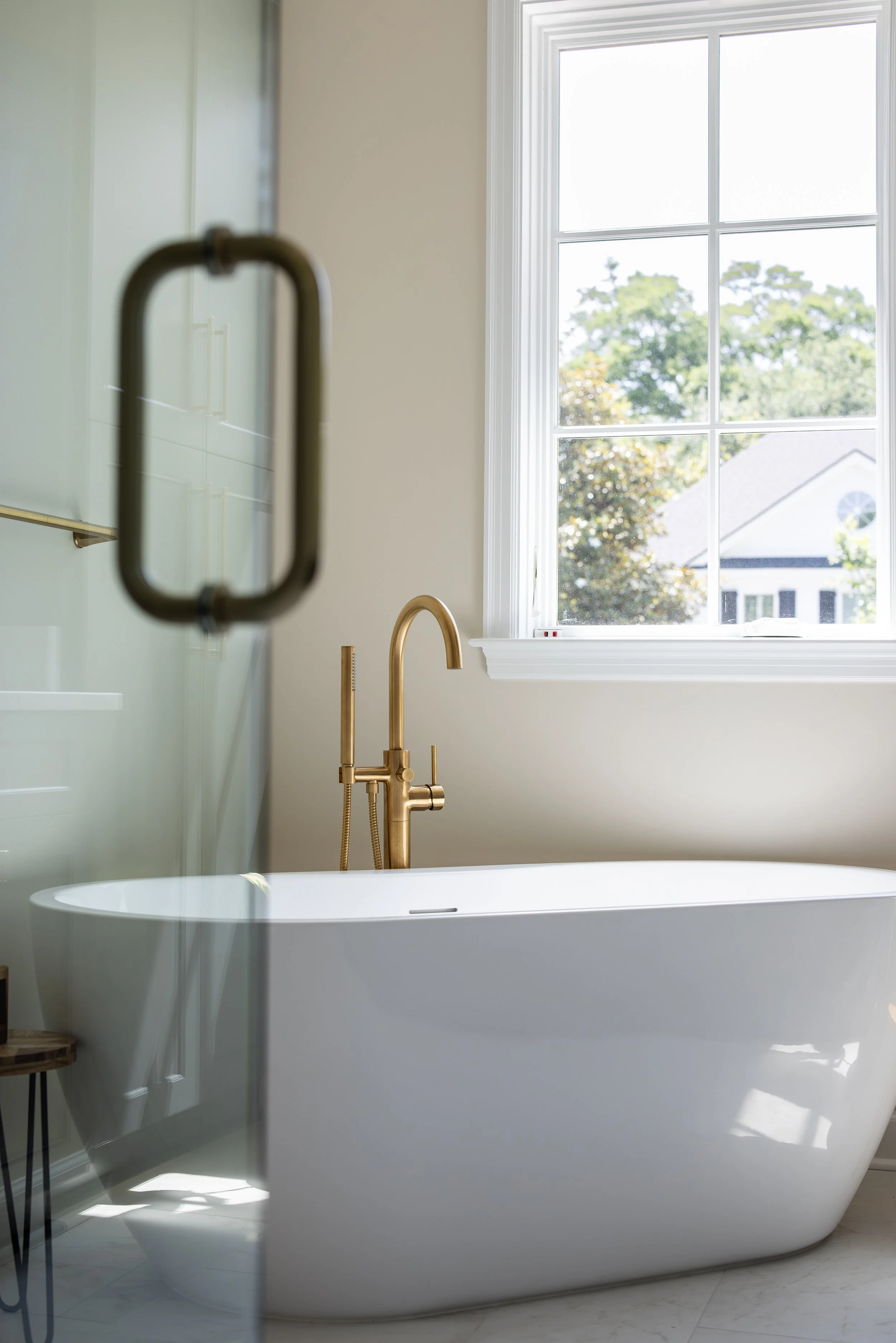 Empty bathroom with a white freestanding bathtub, a gold faucet, a large window showing trees outside, and a partially visible glass shower door.