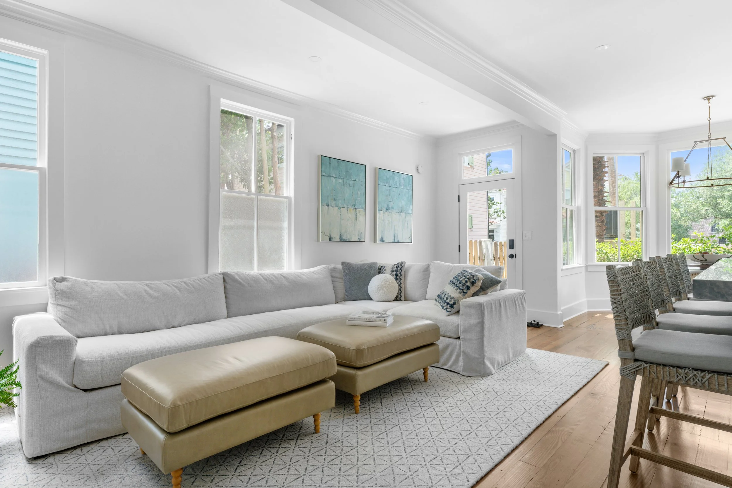 Bright living room with white walls and large windows, featuring a white sofa with assorted cushions, a beige ottoman, a patterned area rug, and dining chairs toward the right. Artwork hangs above the sofa, and a wooden fence is visible outside.