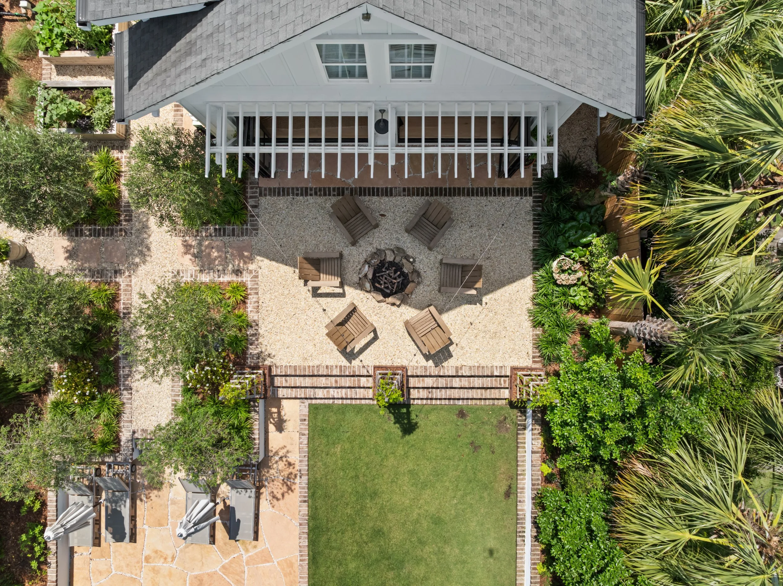 An aerial view of a backyard with a seating area around a fire pit, surrounded by trees and greenery, and an adjacent house with a deck.