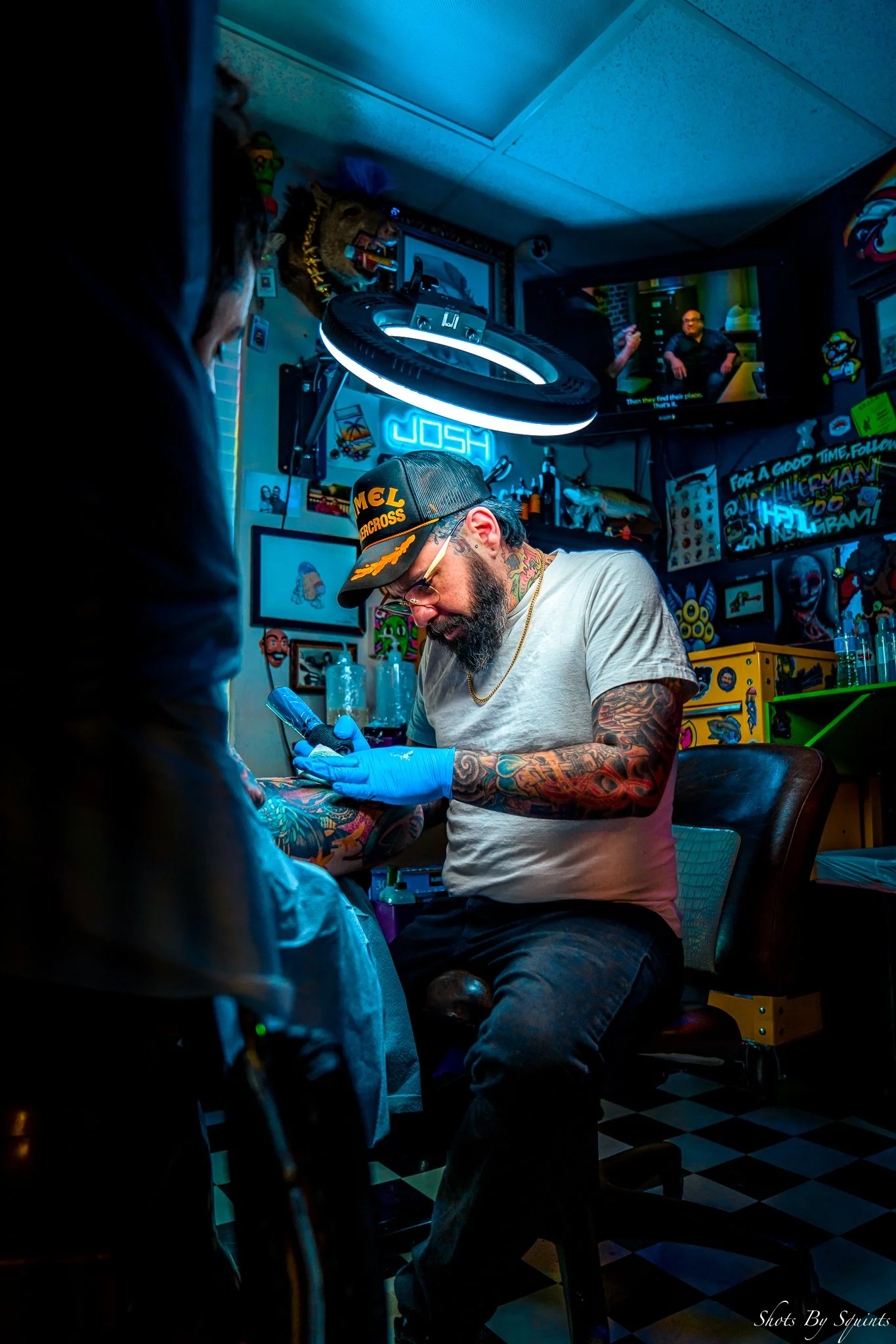 A tattoo artist with glasses, a tattoo on his neck, and colorful tattoos on his arms, wearing a black cap, white t-shirt, and blue gloves, is working on a person's arm in a colorful tattoo studio. The studio has various art pieces and neon signs on t