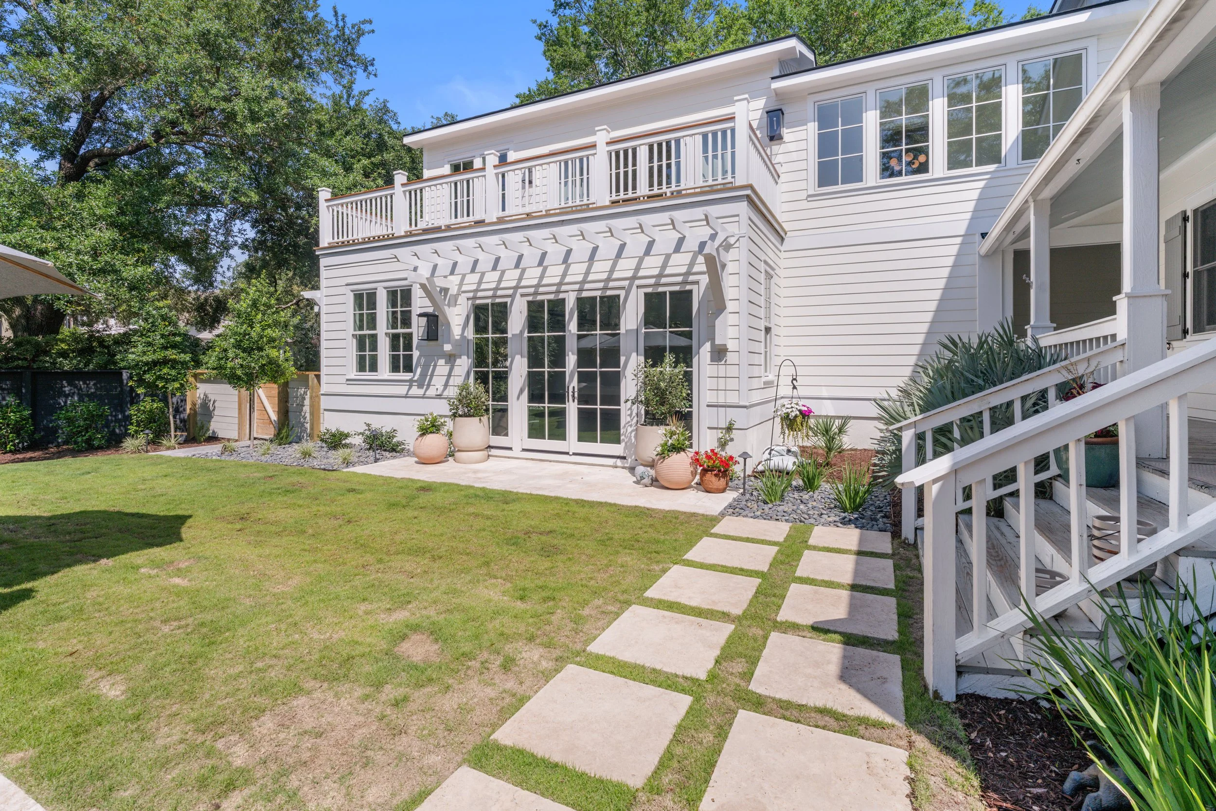 Backyard with a green lawn, stone pathway, potted plants, and a white two-story house with large windows, a balcony, and a porch.