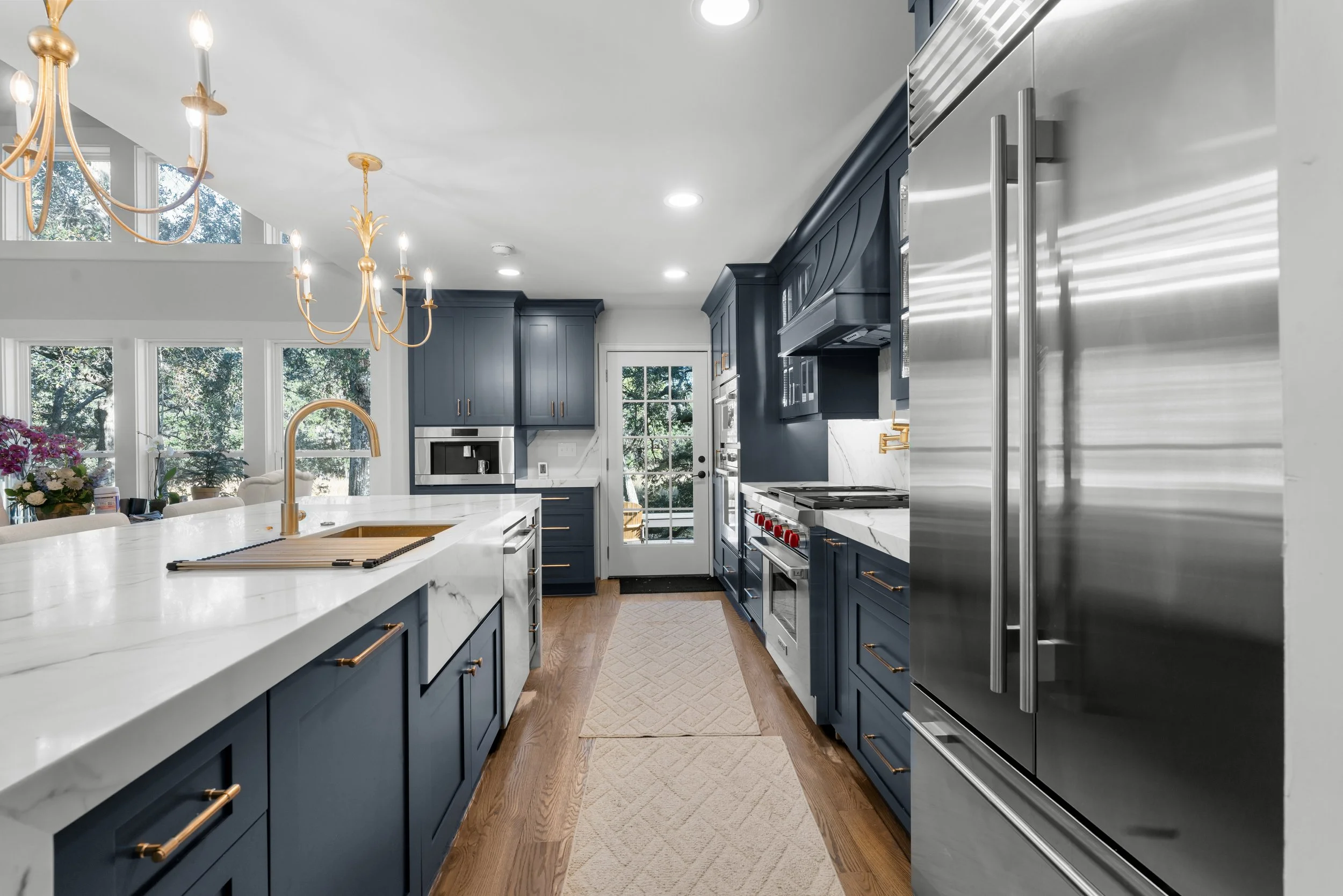 Modern kitchen with navy blue cabinetry, white marble countertops, and gold hardware. Stainless steel refrigerator and appliances, with large windows providing natural light.
