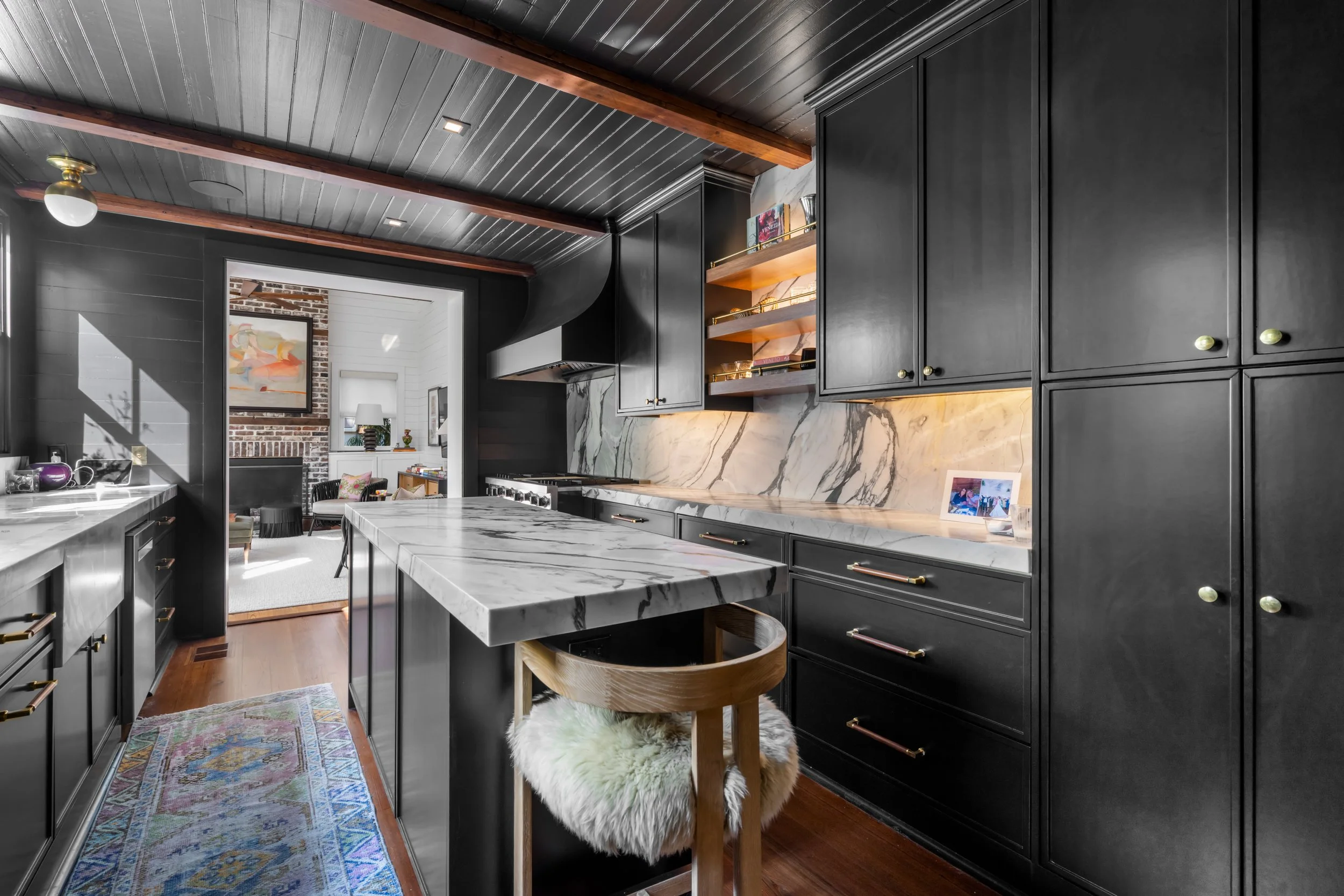 Modern kitchen with black cabinetry, marble countertops, and a marble backsplash; wooden ceiling beams; a kitchen island with a marble top and a wooden stool with a furry cushion; a view into an adjacent sitting area with brick wall, artwork, and sun