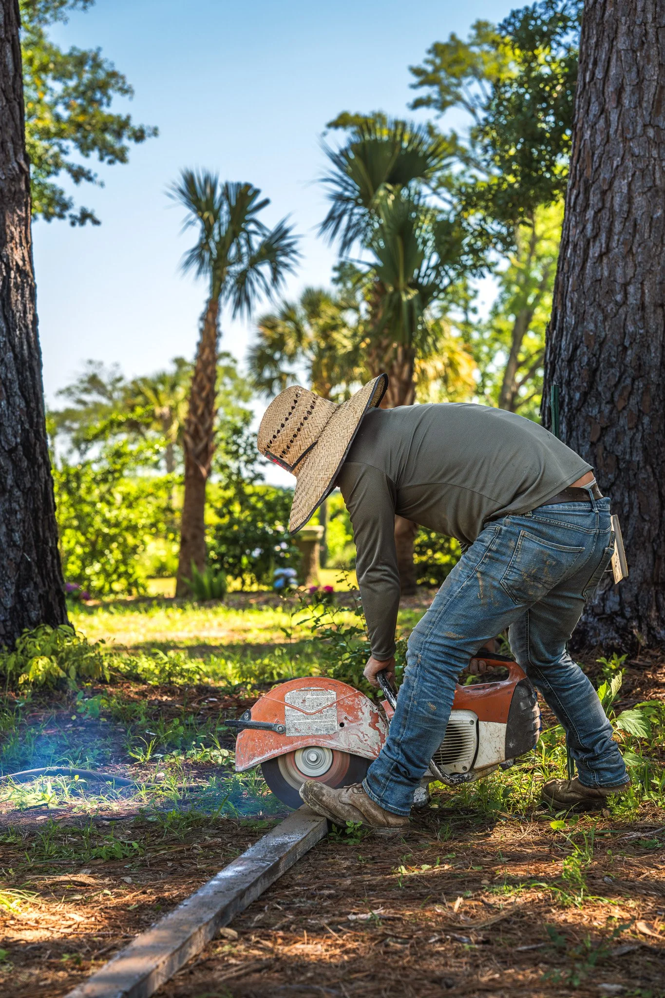 A person using a circular saw to cut a metal rail in a wooded outdoor setting, wearing a straw hat and working between two trees.