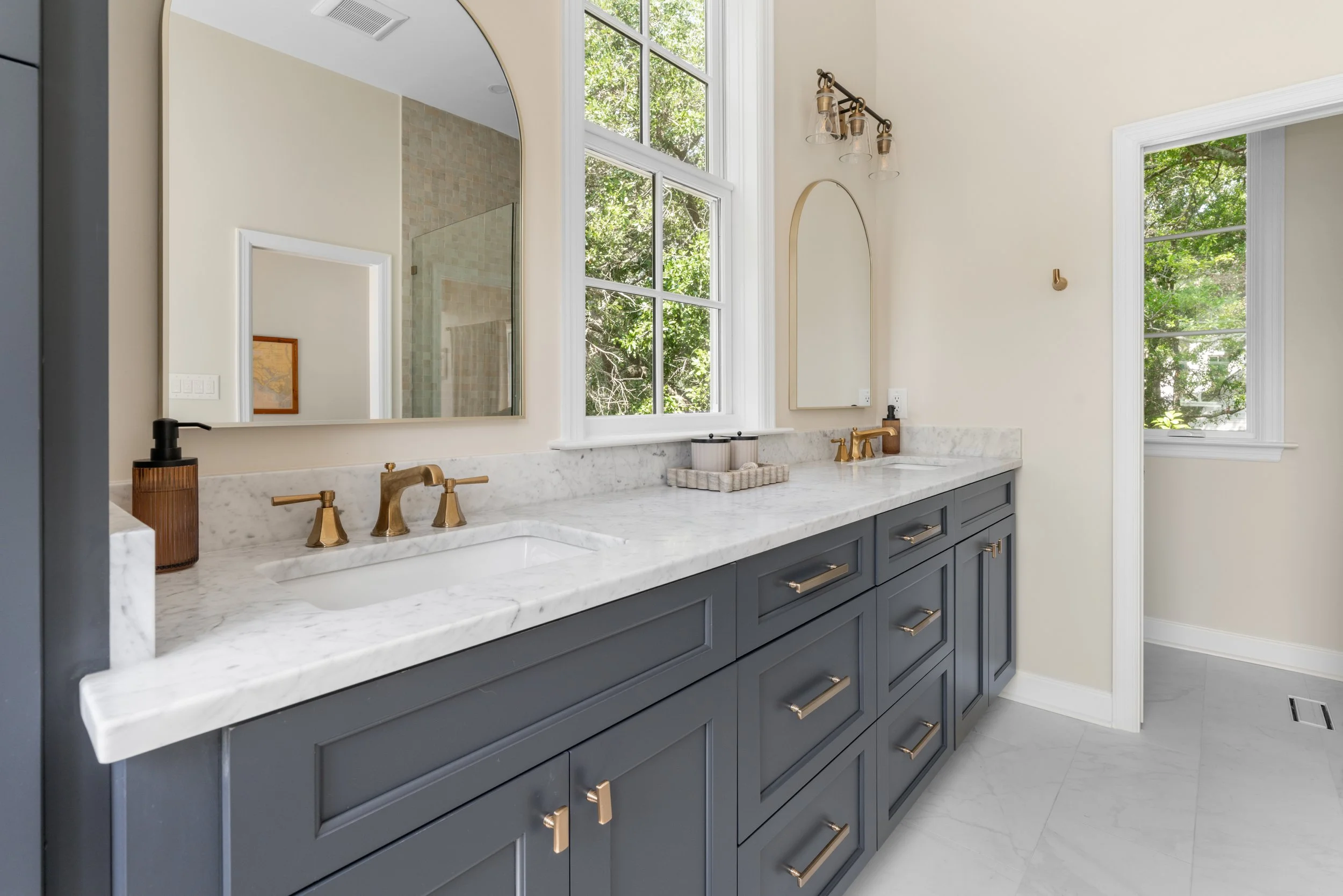 Bathroom vanity with double sinks, marble countertop, navy blue cabinets, gold fixtures, and large mirrors, with windows showing greenery outside.