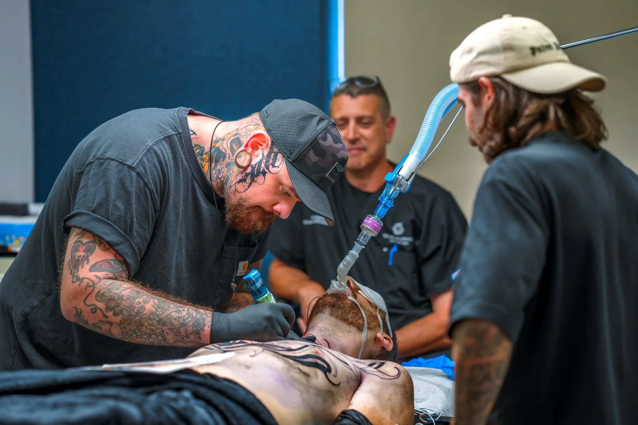 Tattooed man performing a procedure on a patient with personal protective equipment, while two other men observe in a medical setting.
