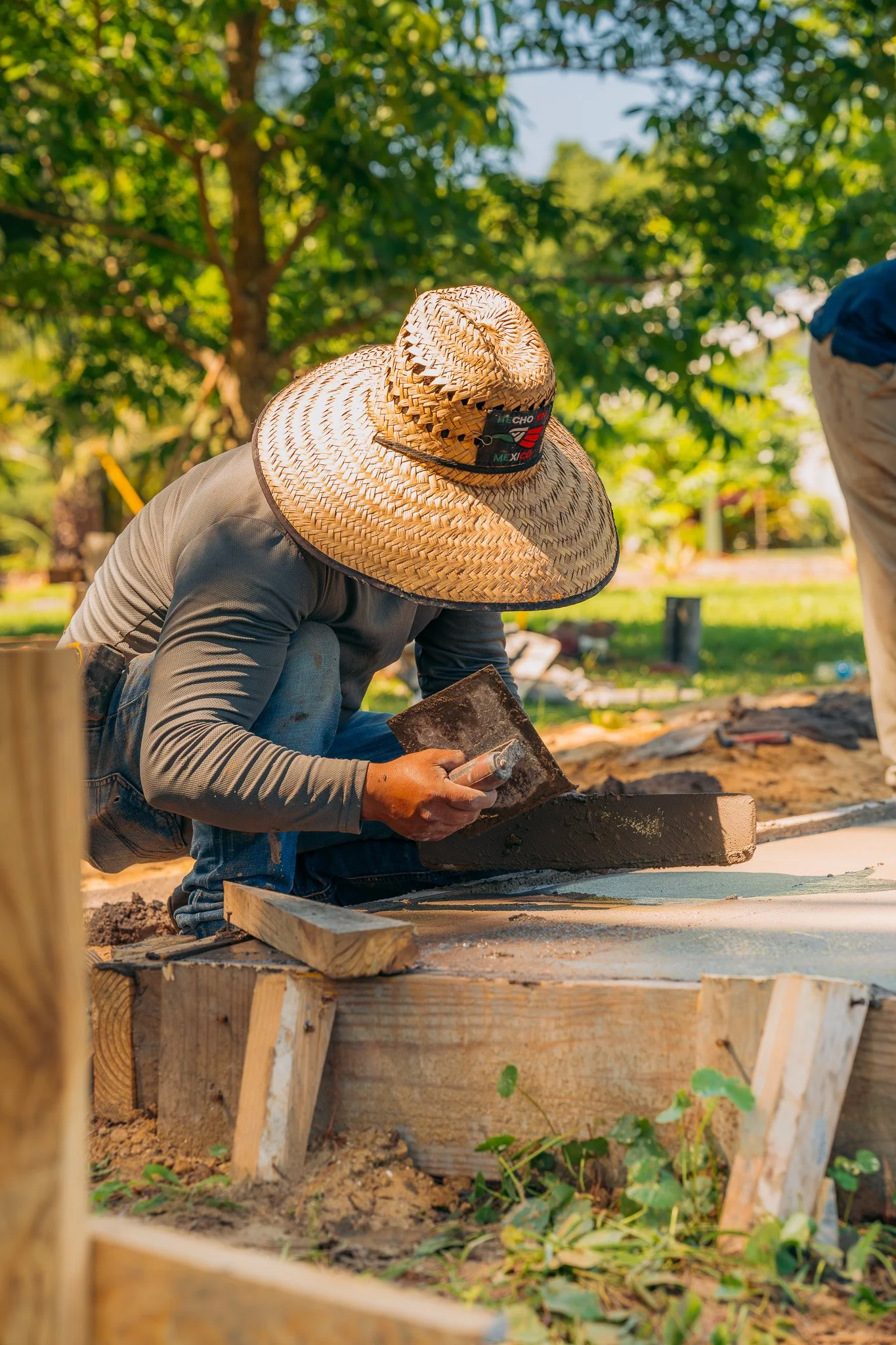 A man wearing a wide-brimmed straw hat working on a construction site outdoors, kneeling down and working with concrete or mortar on a wooden form.
