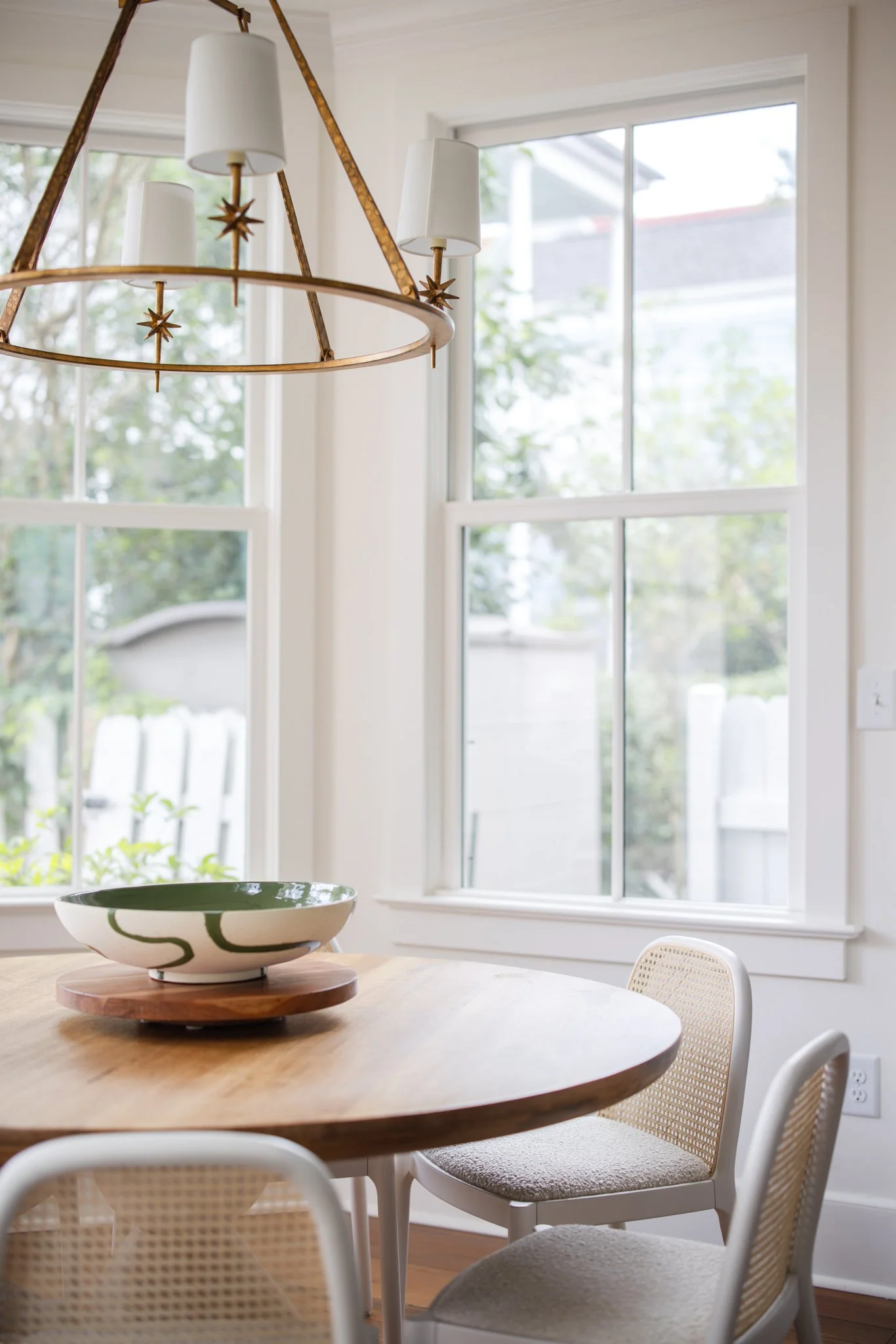 A dining area with a round wooden table, surrounded by chairs with woven backs and cushioned seats, a large decorative ceramic bowl on a wooden stand, and a gold-colored chandelier hanging above. Large windows let in natural light and offer a view of