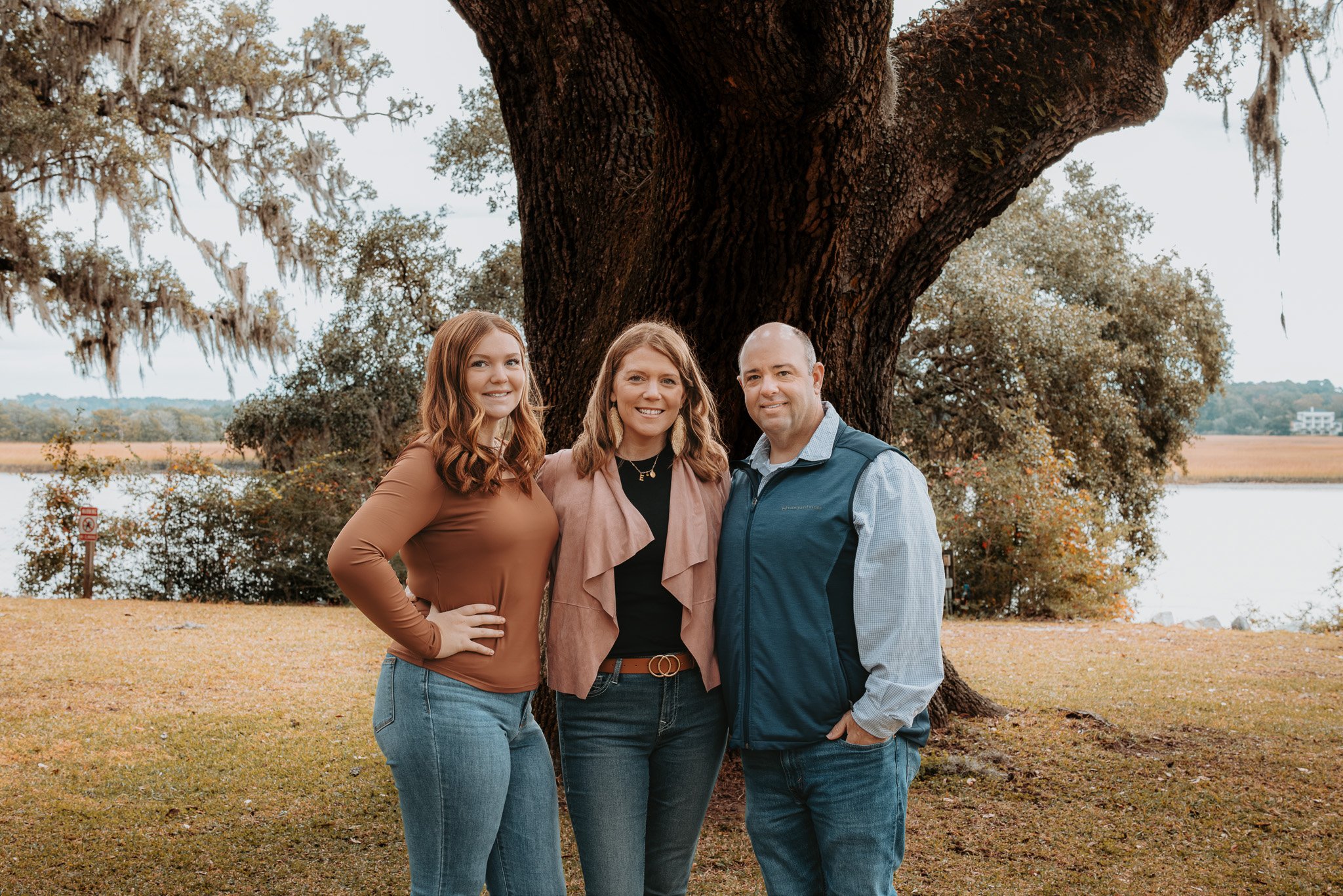 Three people standing outdoors in front of a large tree with Spanish moss hanging from the branches. They are near a body of water with a landscape of fields and trees in the background. They are smiling and posing for the photo.