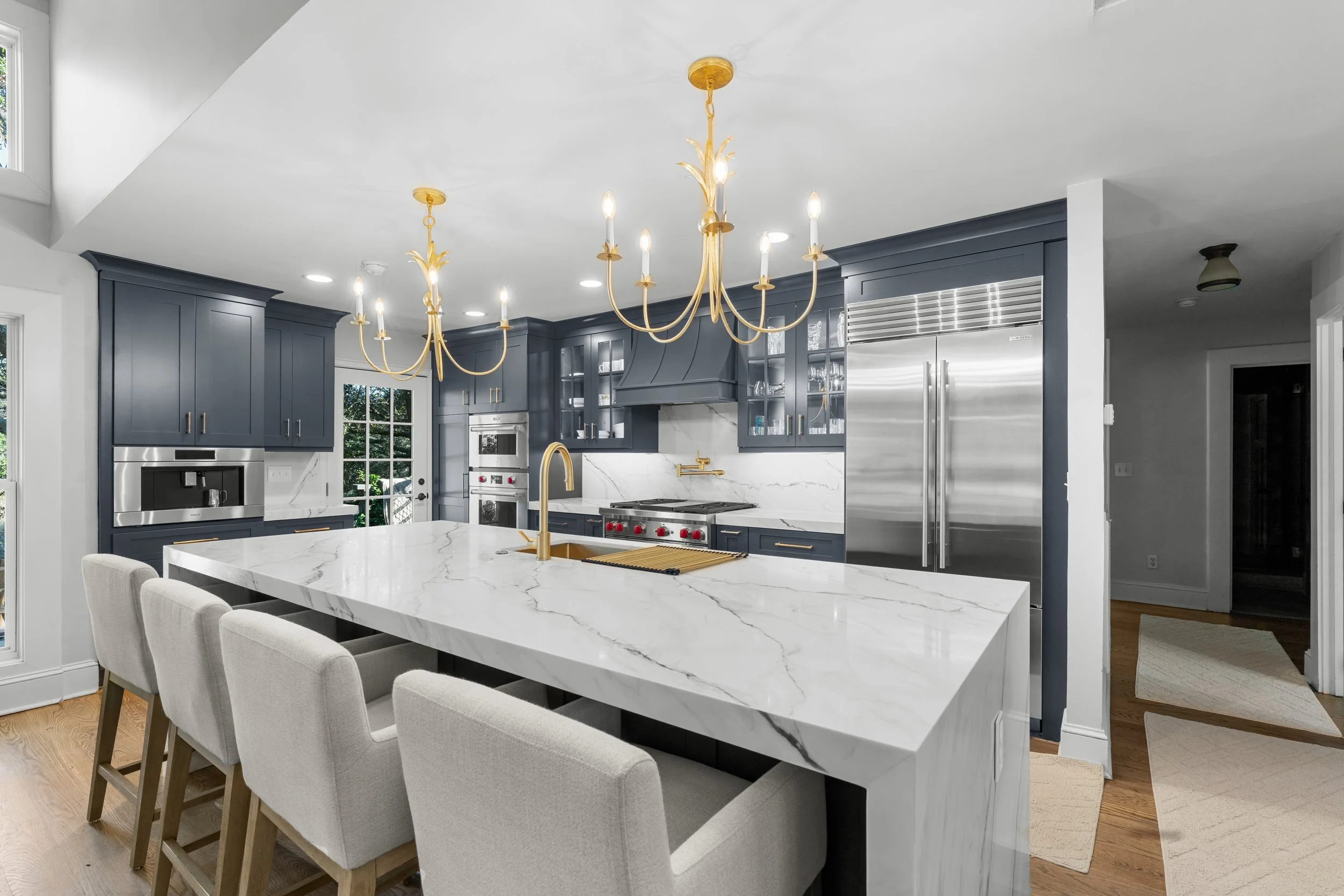 Modern kitchen with white marble island, navy blue cabinets, stainless steel appliances, and gold fixtures, including chandeliers and a faucet.