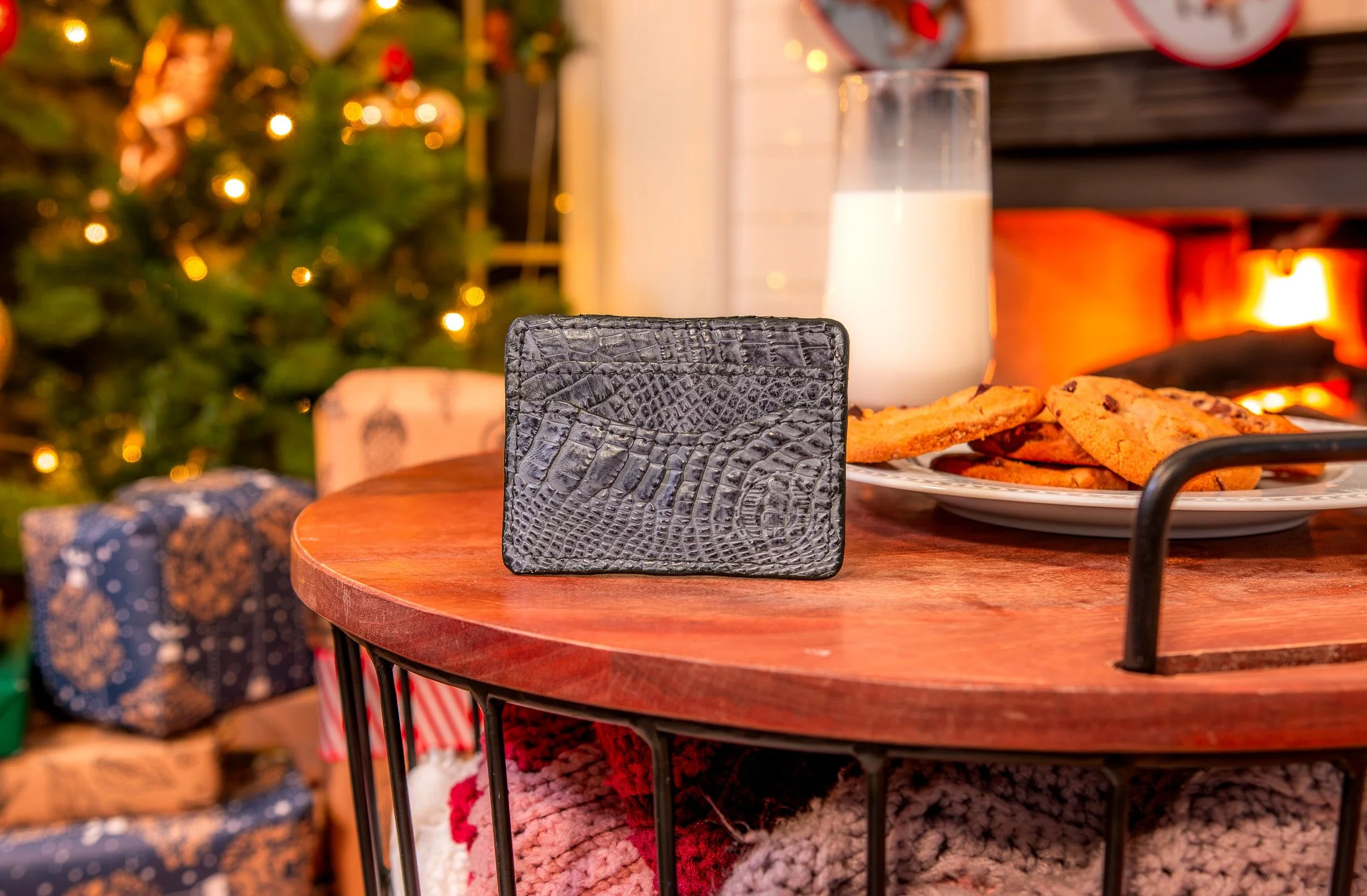 A black textured wallet, a glass of milk, and a plate of chocolate chip cookies on a round wooden table, with Christmas presents and a decorated Christmas tree in the background.
