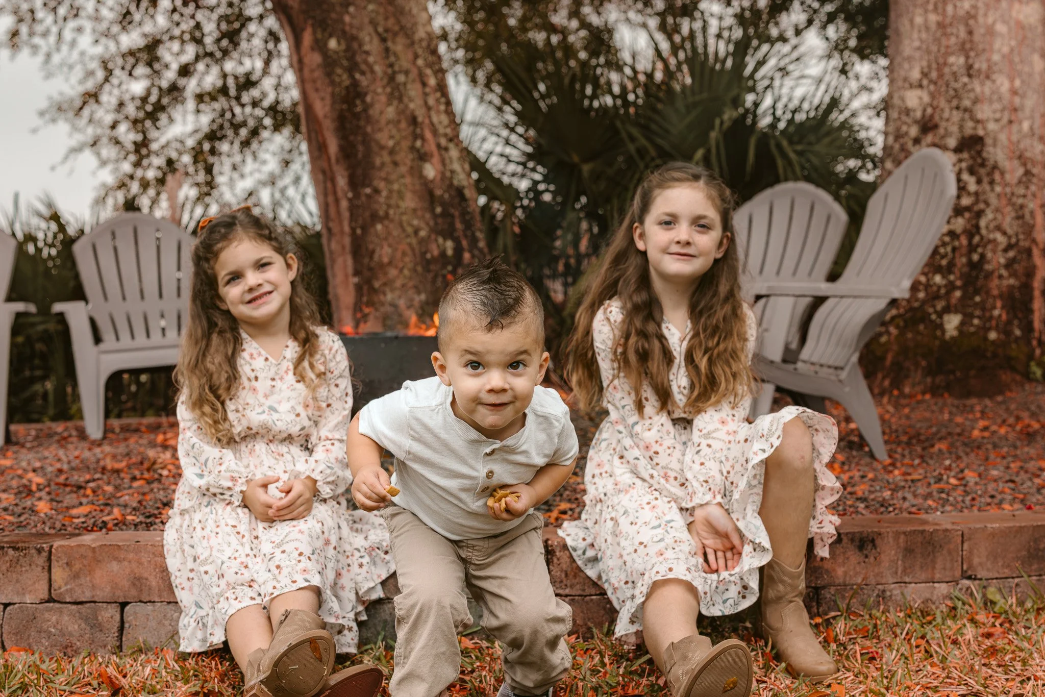 Three children, two girls and one boy, sitting outdoors on a brick wall in a wooded area with large trees, fallen leaves, and Adirondack chairs, near a fire pit.