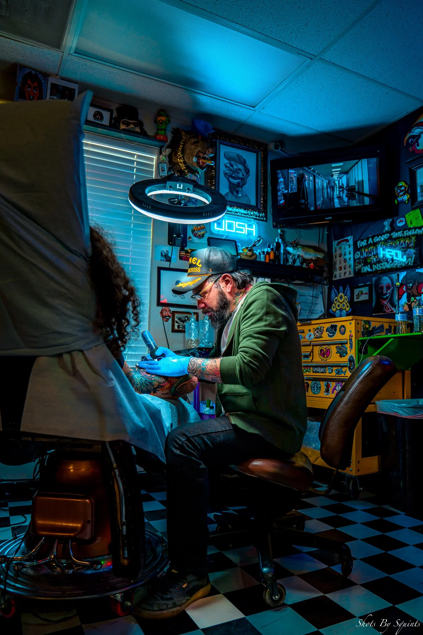 A tattoo artist working on a client's arm in a colorful, decorated tattoo studio with neon signs and artwork.