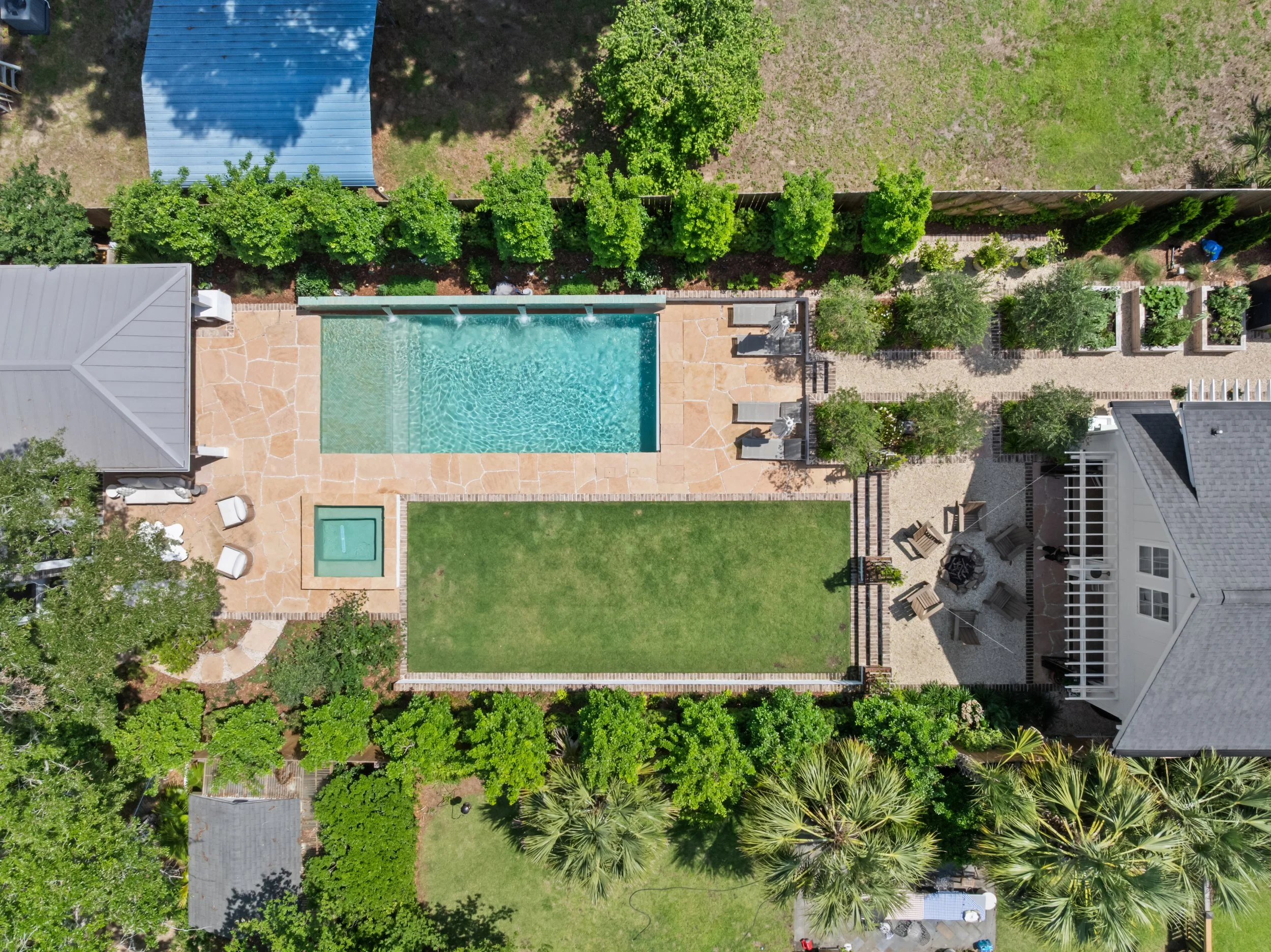 Aerial view of a backyard with a swimming pool, hot tub, grassy area, patio with chairs and fire pit, surrounded by green trees and shrubs.