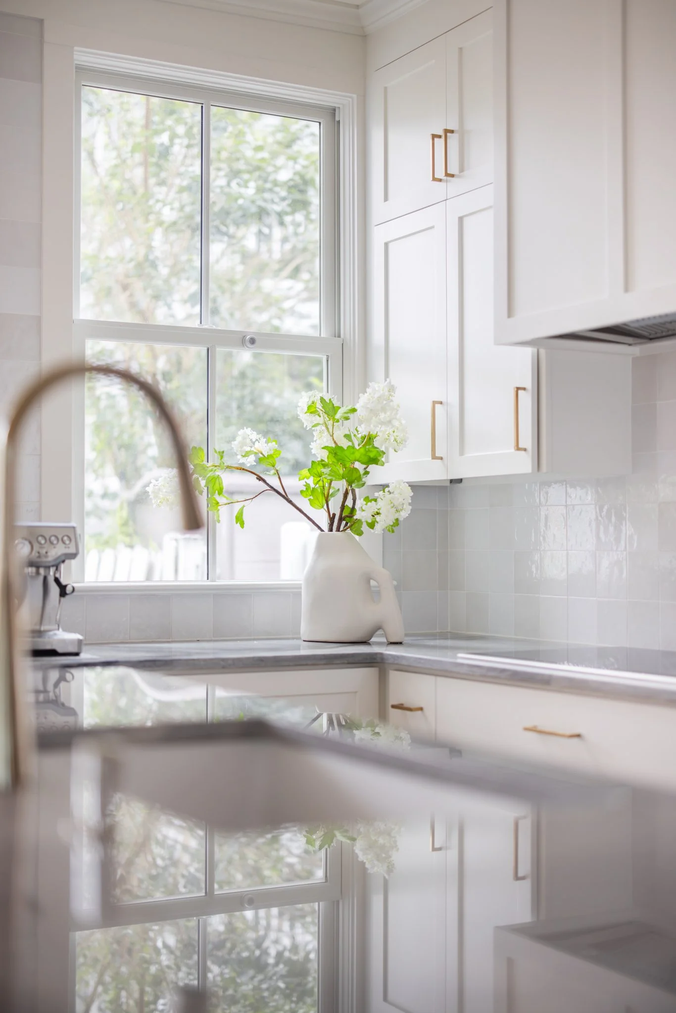 White kitchen with a large window, white cabinetry with gold handles, and a white vase with green and white flowers on the countertop.