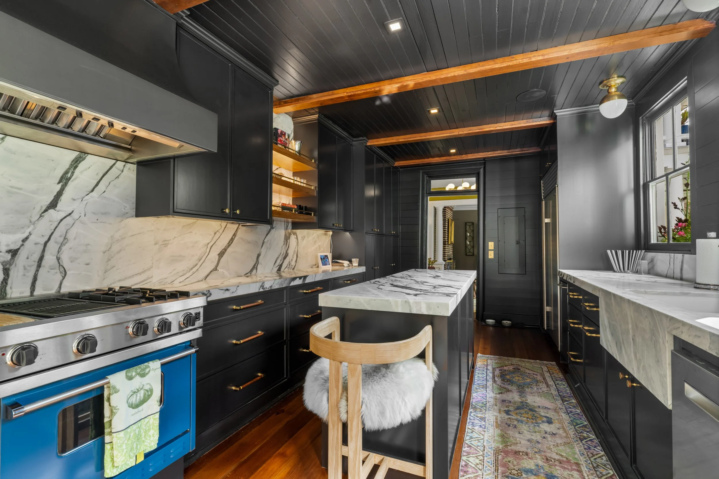 Modern kitchen with black cabinets, white marble countertops and backsplash, a blue oven with a green dish towel, a wooden stool with a fluffy white cushion, and large windows with a view of pink flowers outside.