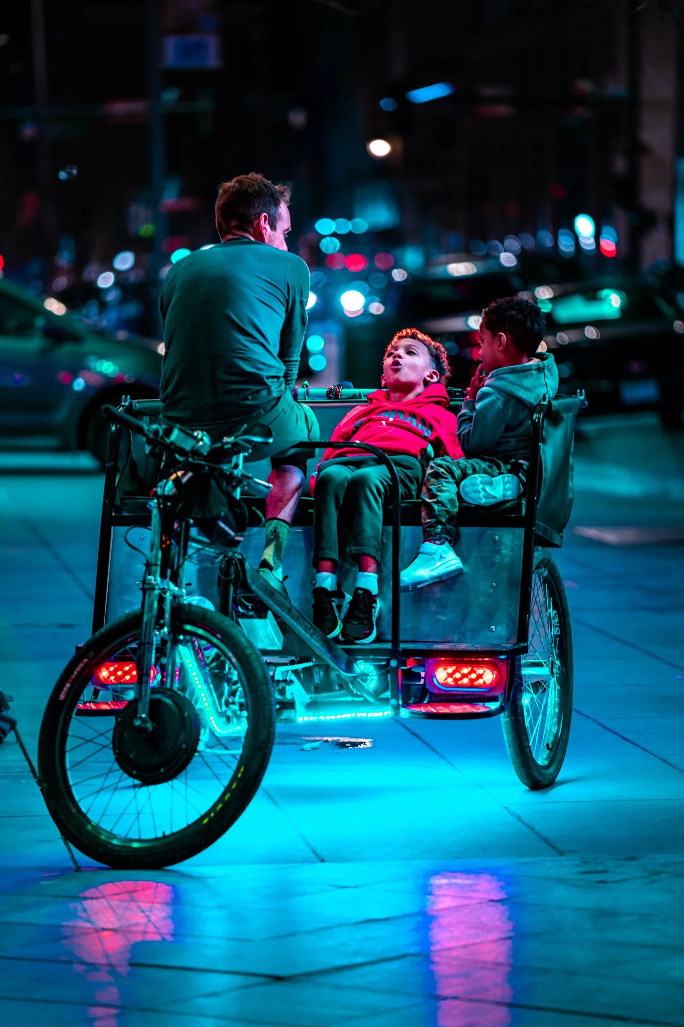 A man on a tricycle with two children at night, surrounded by city lights and blurred cars.