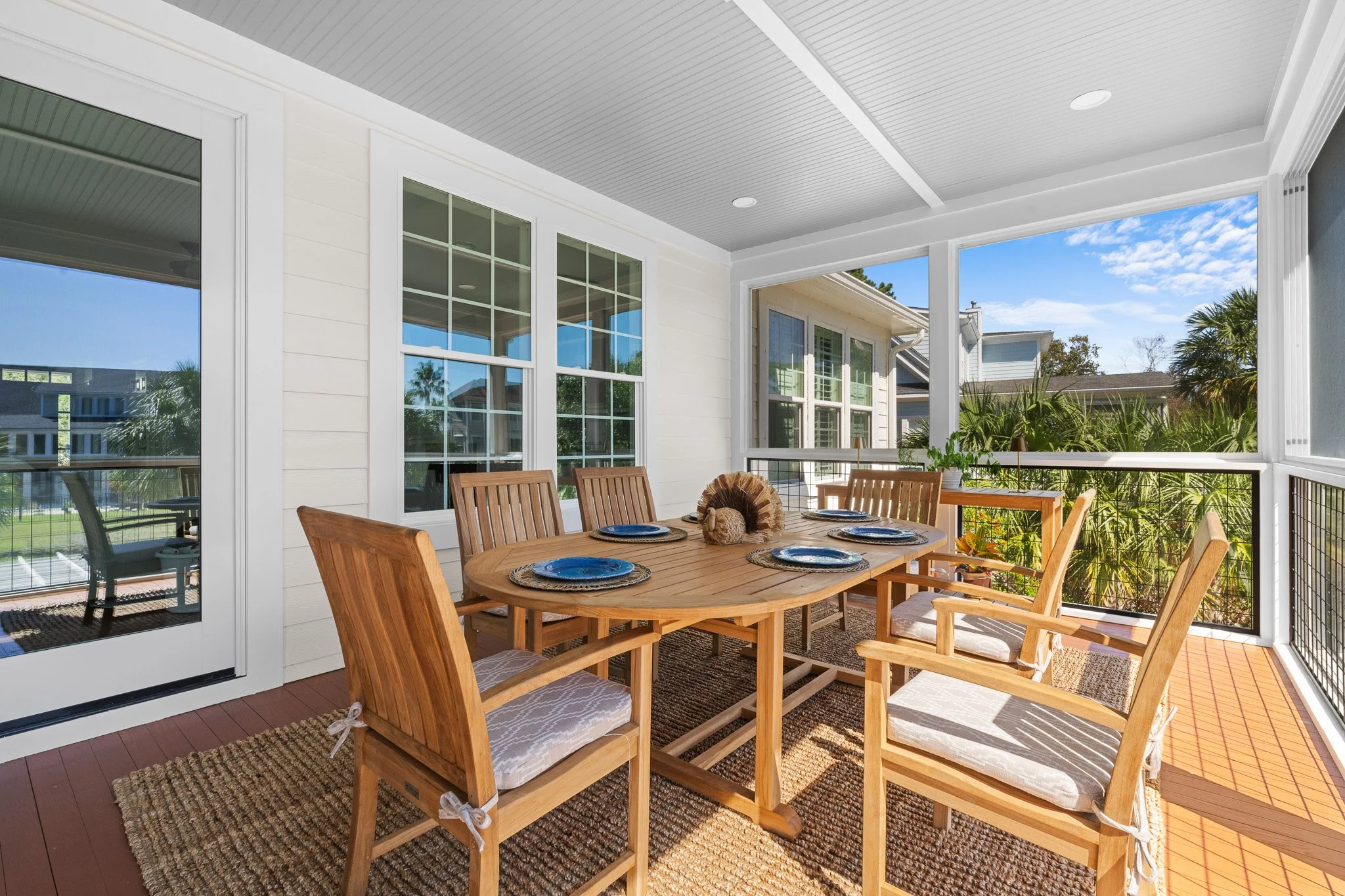 A screened porch with a round wooden dining table set with blue plates and eight wooden chairs with cushions, overlooking a sunny yard with greenery and neighboring houses.