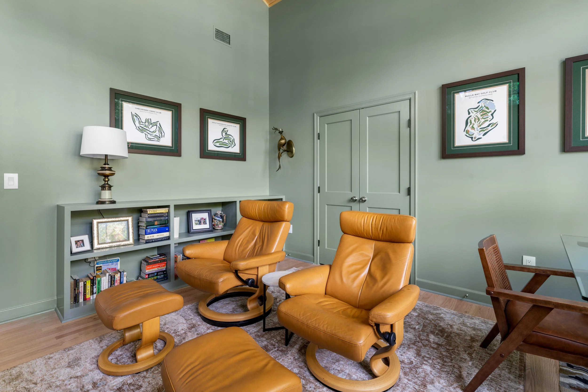 A cozy living room with light green walls, featuring two tan leather chairs, a matching ottoman, a small side table, a bookshelf with books and framed photos, three framed golf course maps on the wall, a white lamp, a piece of wall art, and a wooden 