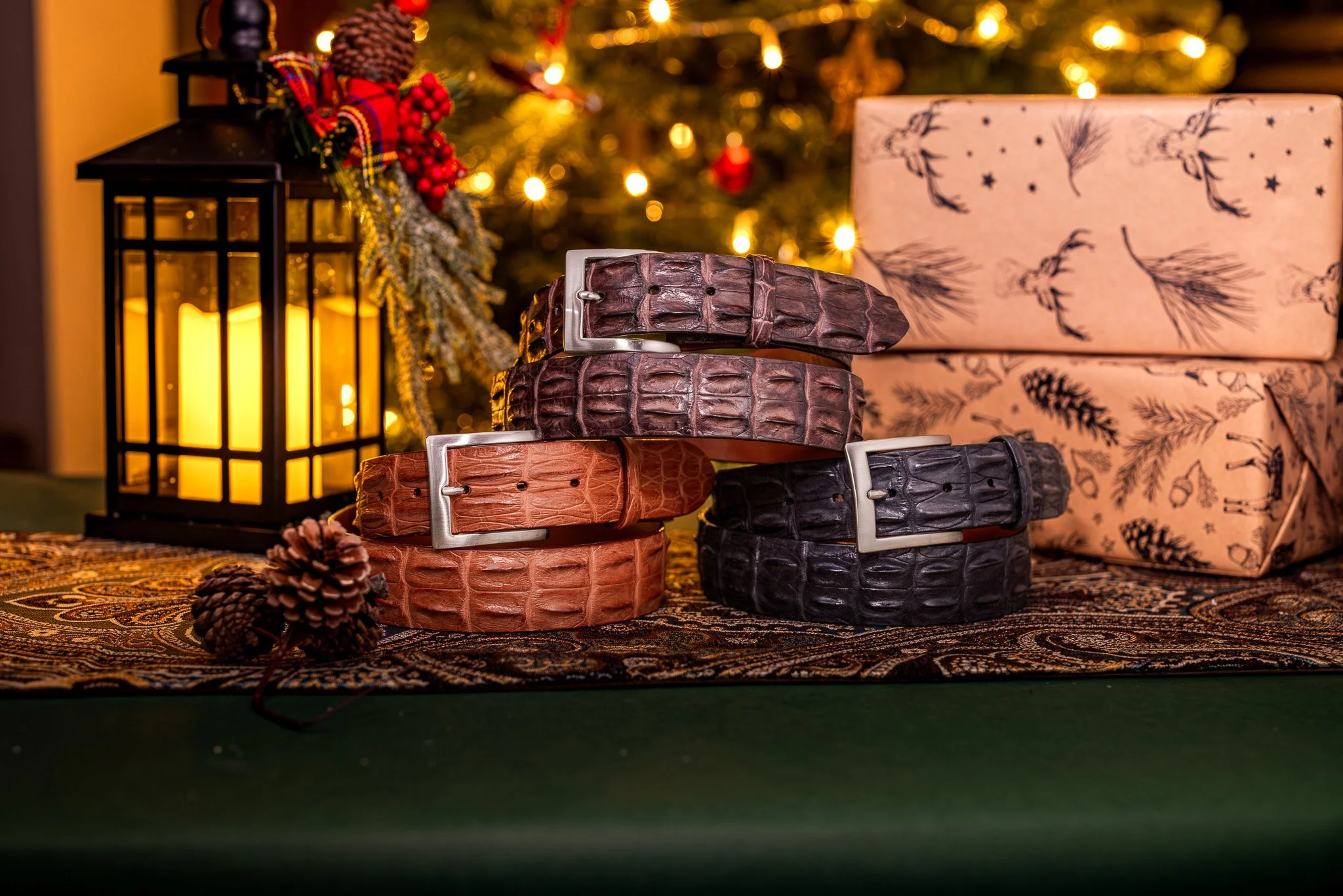 Four leather belts with crocodile texture in black, dark brown, tan, and reddish-brown, arranged in front of a Christmas tree with blurred lights, next to a black lantern with candles, pinecones, and wrapped gifts with holiday-patterned paper.