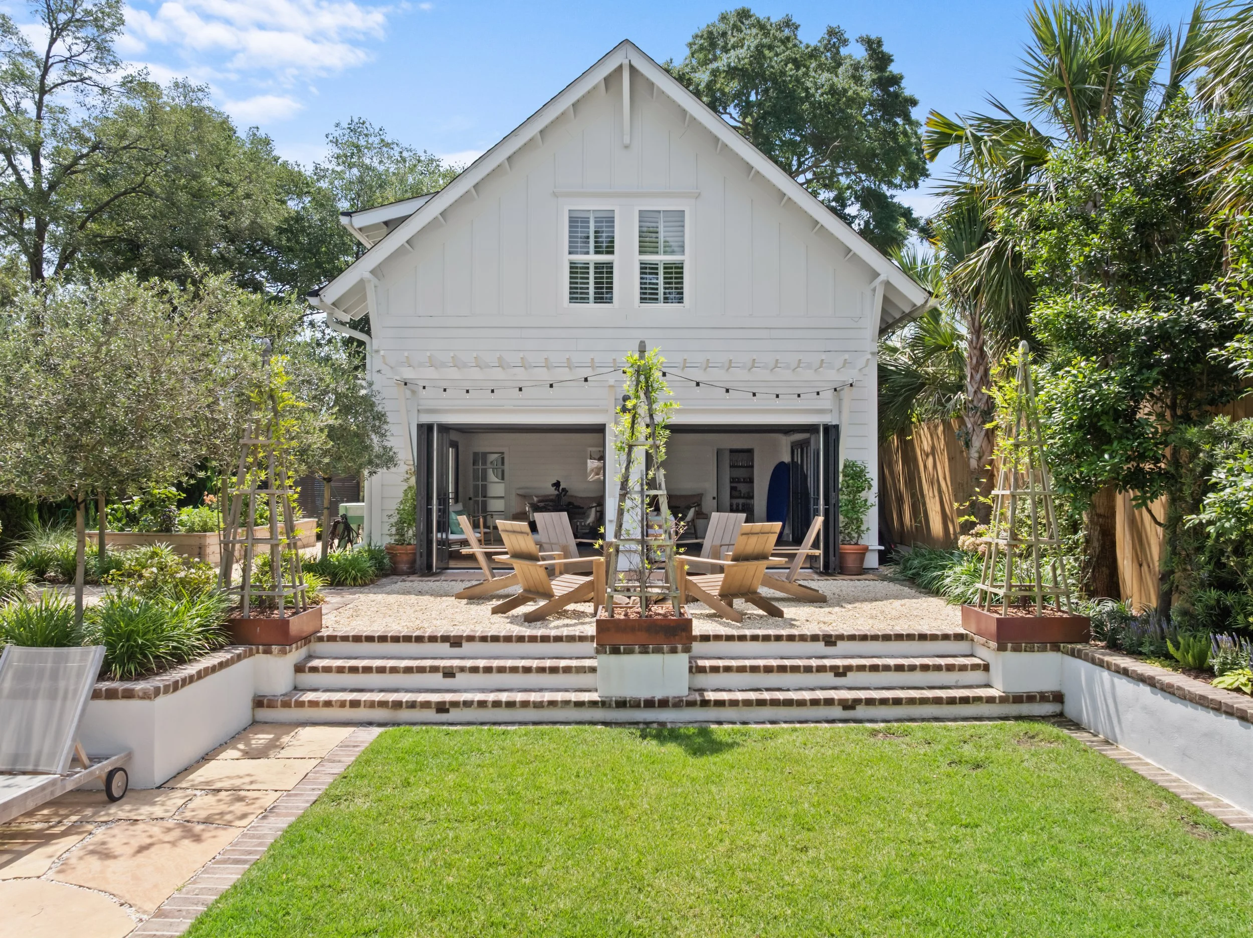 A backyard with green grass, brick steps leading up to a white cottage-style house with open garage doors, outdoor seating, potted plants, trees, and string lights.