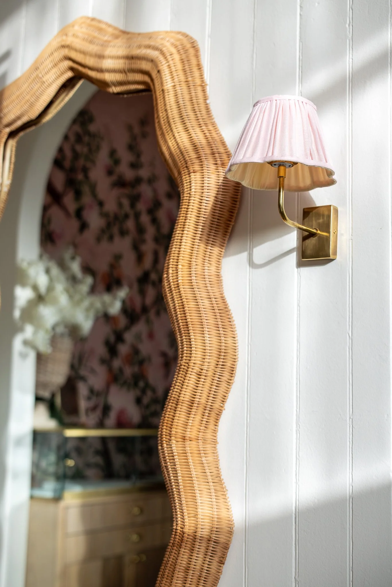 A close-up of a wavy rattan mirror frame mounted on a white wall, with a brass wall sconce and pink pleated lampshade visible nearby.