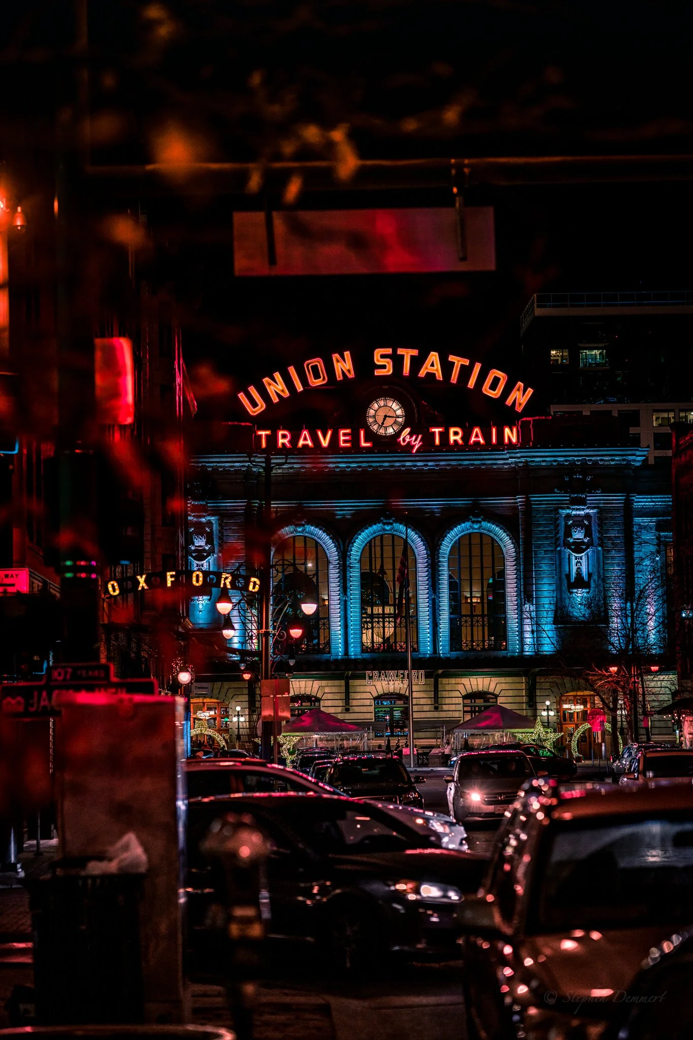 Night view of Union Station with neon lights, the sign reads 'Union Station Travel by Train'. Cars parked in front of the station and illuminated street lamps are visible.