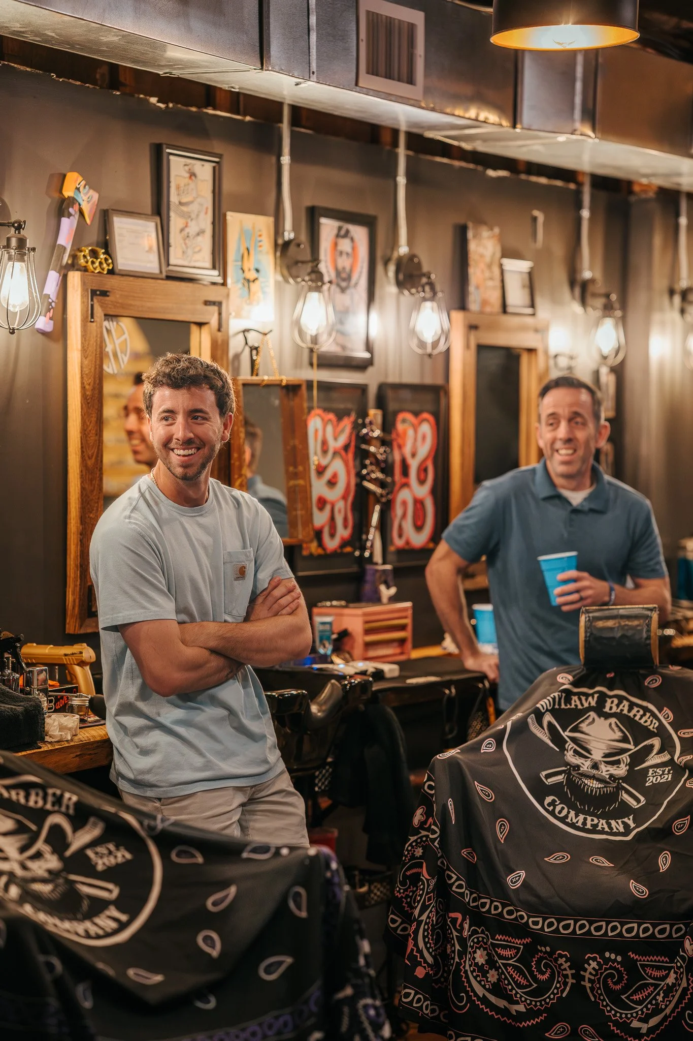 Two men inside a barbershop, smiling and holding blue cups, with barber capes in the foreground and art on the wall behind them.