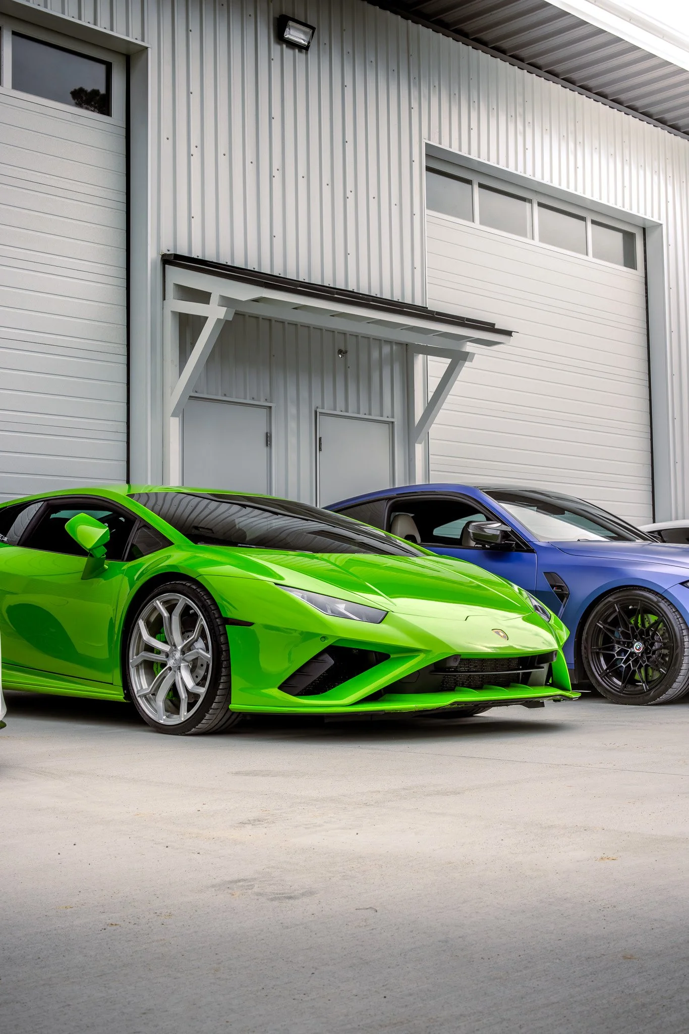 A bright green Lamborghini and a blue sports car parked outside a white metal industrial building with a garage door.