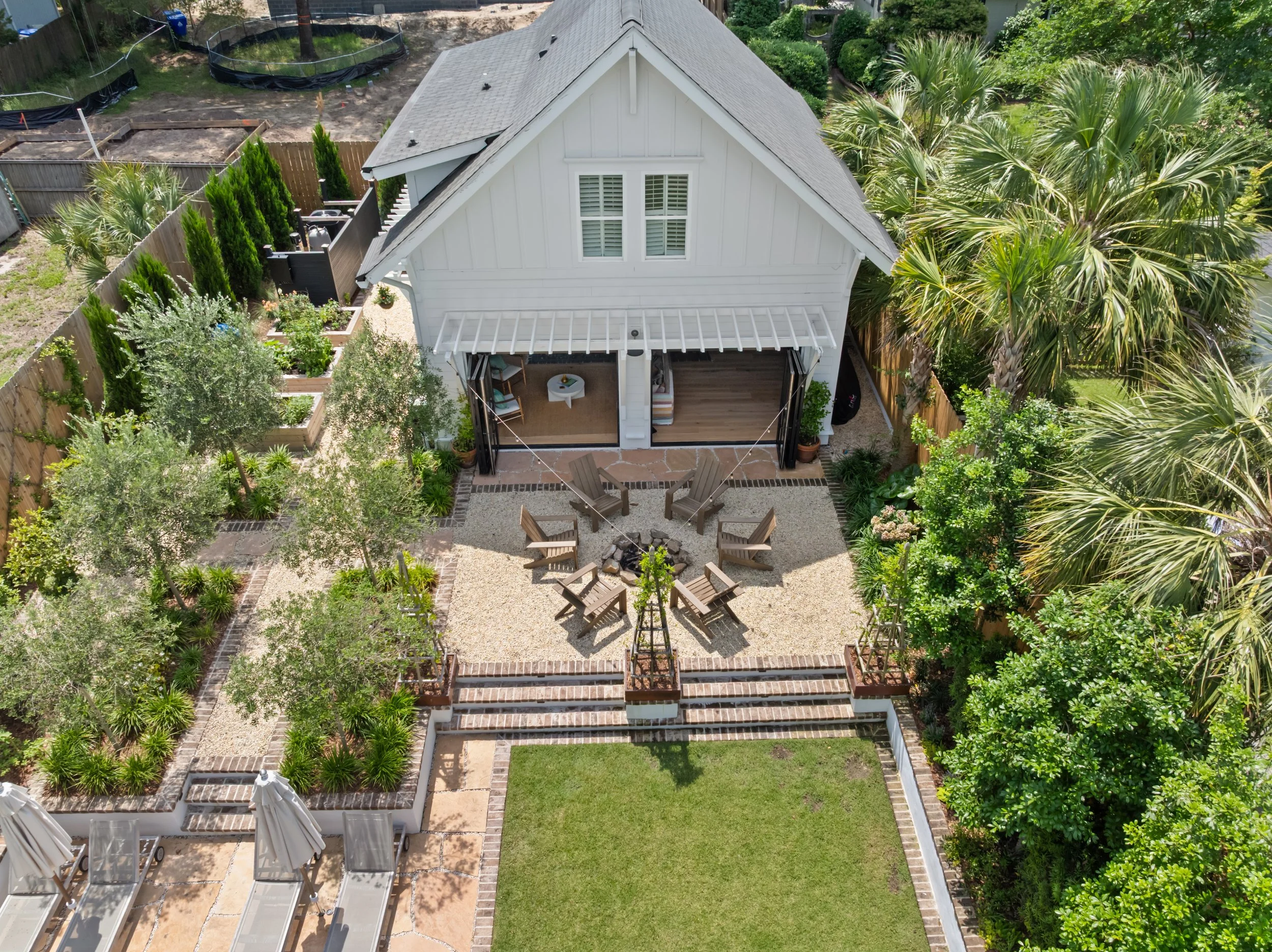 Aerial view of a backyard with a white house, a fire pit circle with wooden chairs, lush greenery including palm trees, and garden beds.