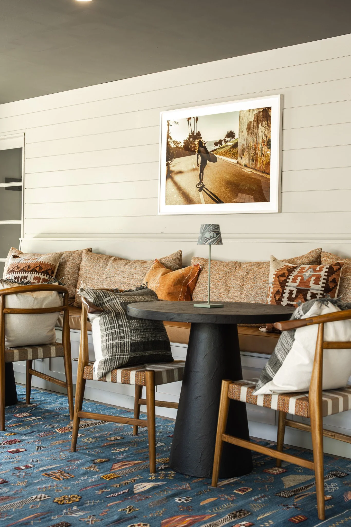 Living room with beige sofa, patterned pillows, wooden chairs, black table with a lamp, framed photo of a skateboarder on the wall, and a blue patterned rug.
