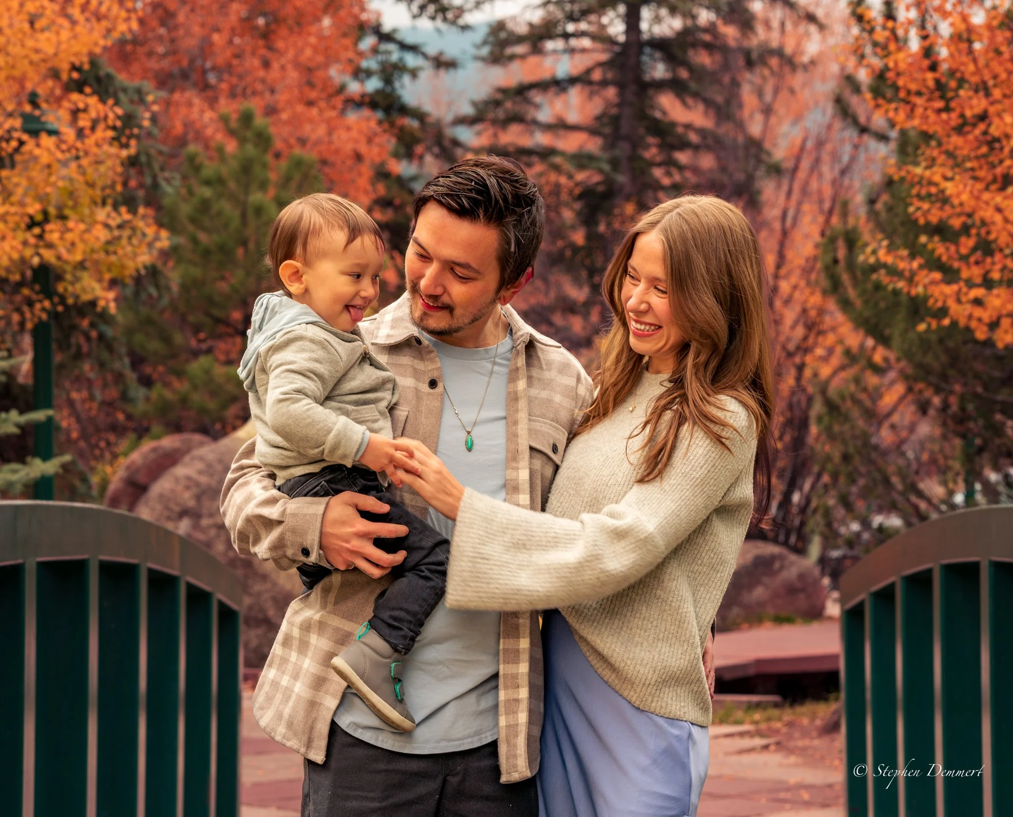 A family of three enjoying a fall day outdoors, with colorful autumn trees in the background. The father is holding a young child, and the mother is reaching to touch the child's hand, all smiling.
