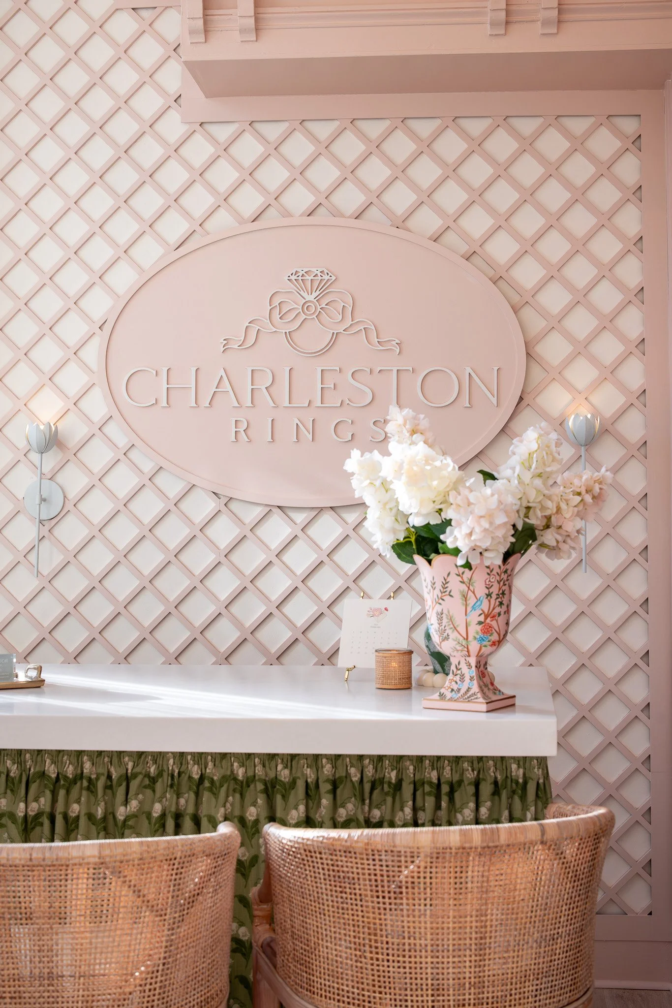 Interior of a jewelry store with a pink lattice wall, a sign saying 'Charleston Rings' with a diamond and bow logo, a white desk with a pink floral vase filled with white flowers, and wicker chairs in the foreground.