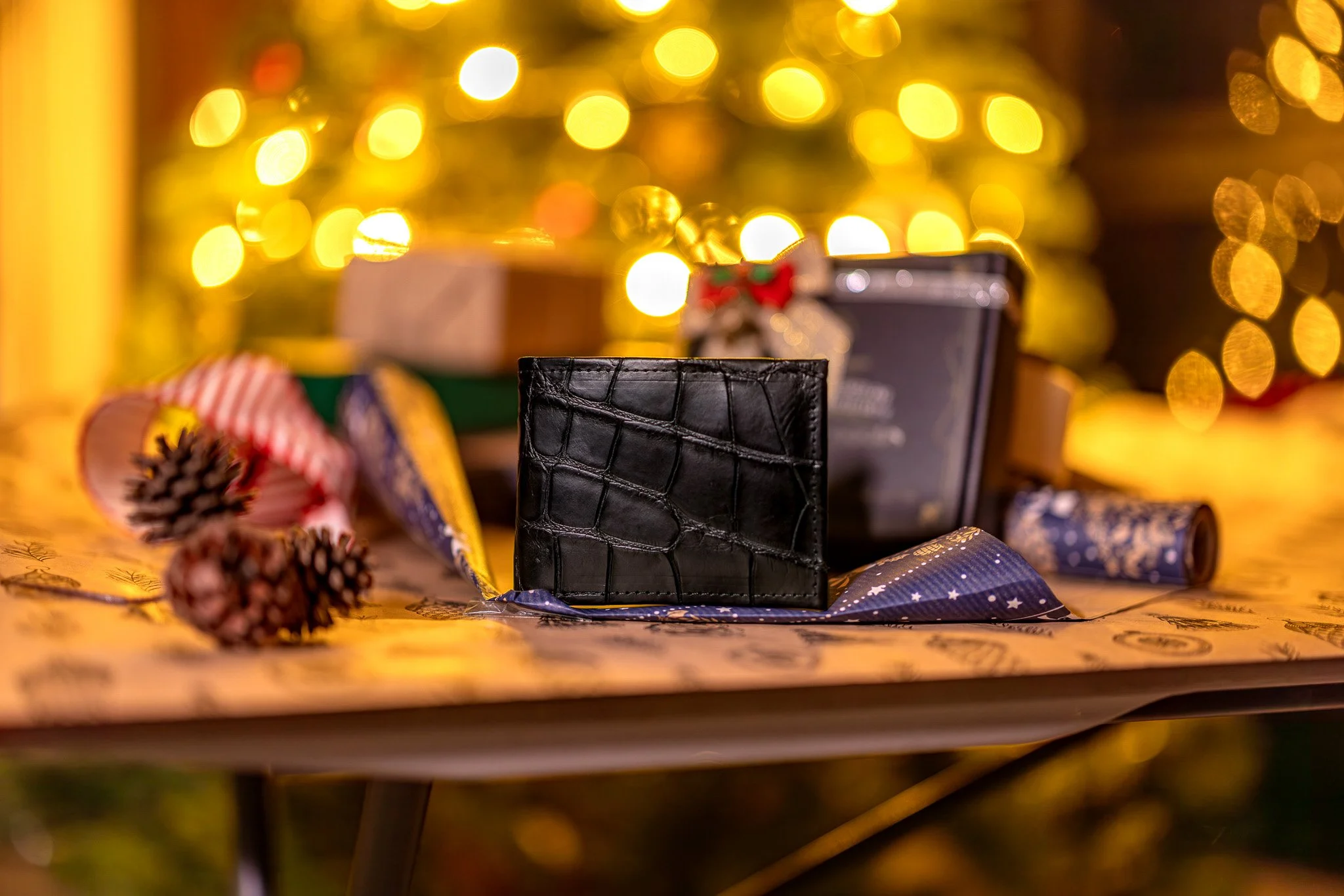 Black wallet with a textured pattern on a table, surrounded by pinecones, ribbon, and gift boxes, with Christmas lights in the background.