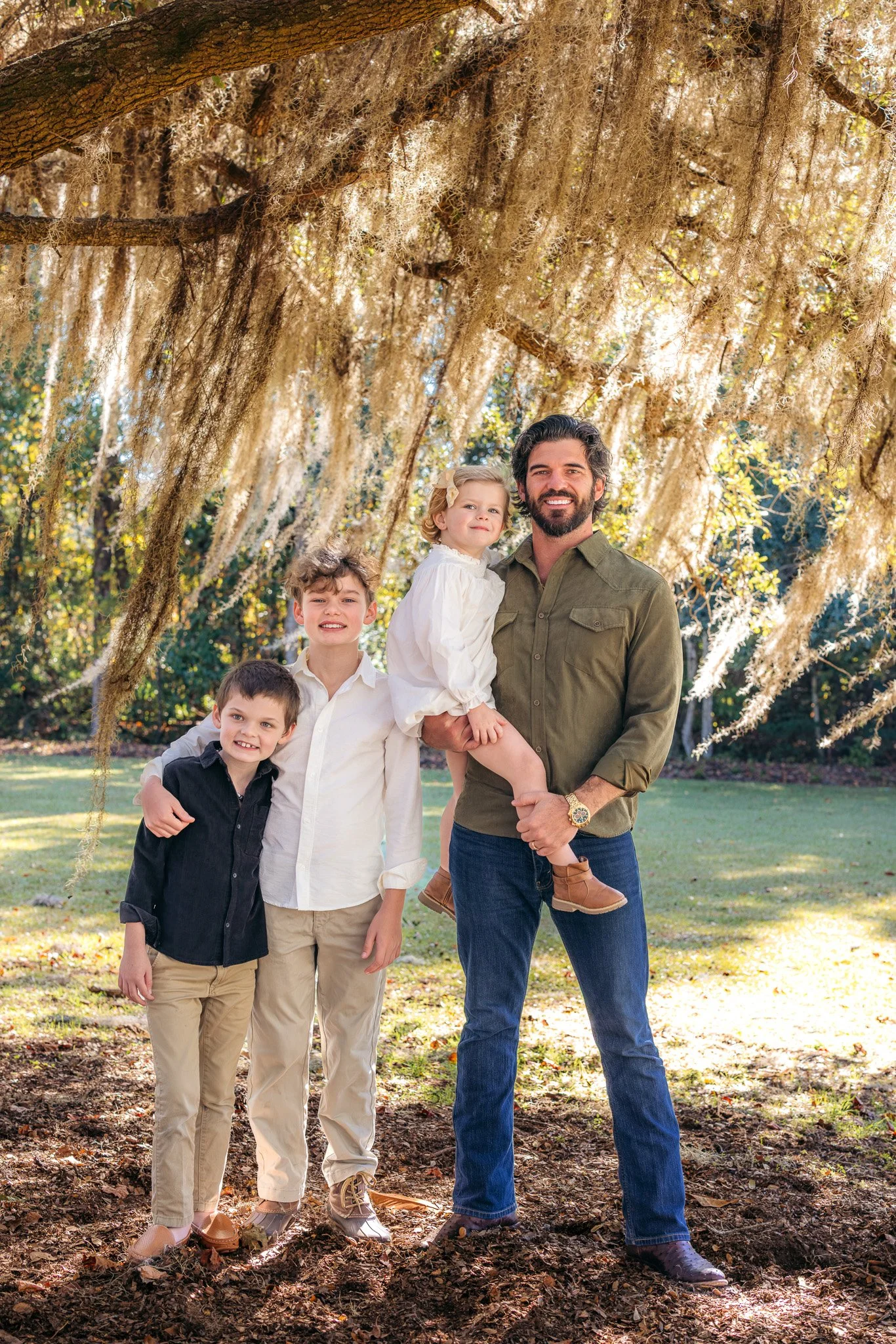 A family of four, including a man, woman, and two children, posing outdoors under a large tree with hanging moss. The family is smiling and dressed casually, with trees and grass in the background.