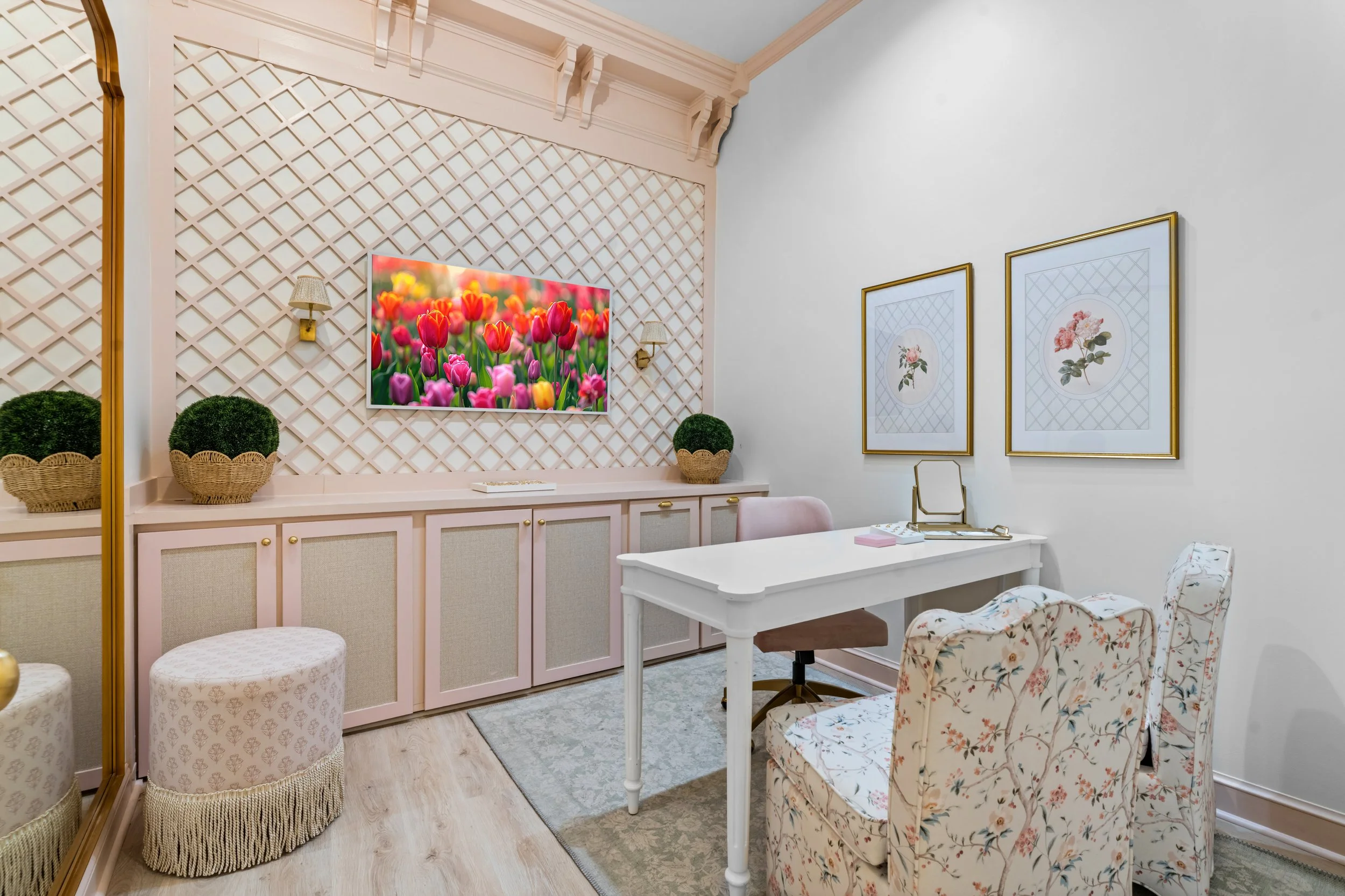 A cozy room with a white desk, floral-patterned chairs, framed botanical prints on the wall, a wall-mounted TV displaying colorful tulips, and decorative plants on the countertop.