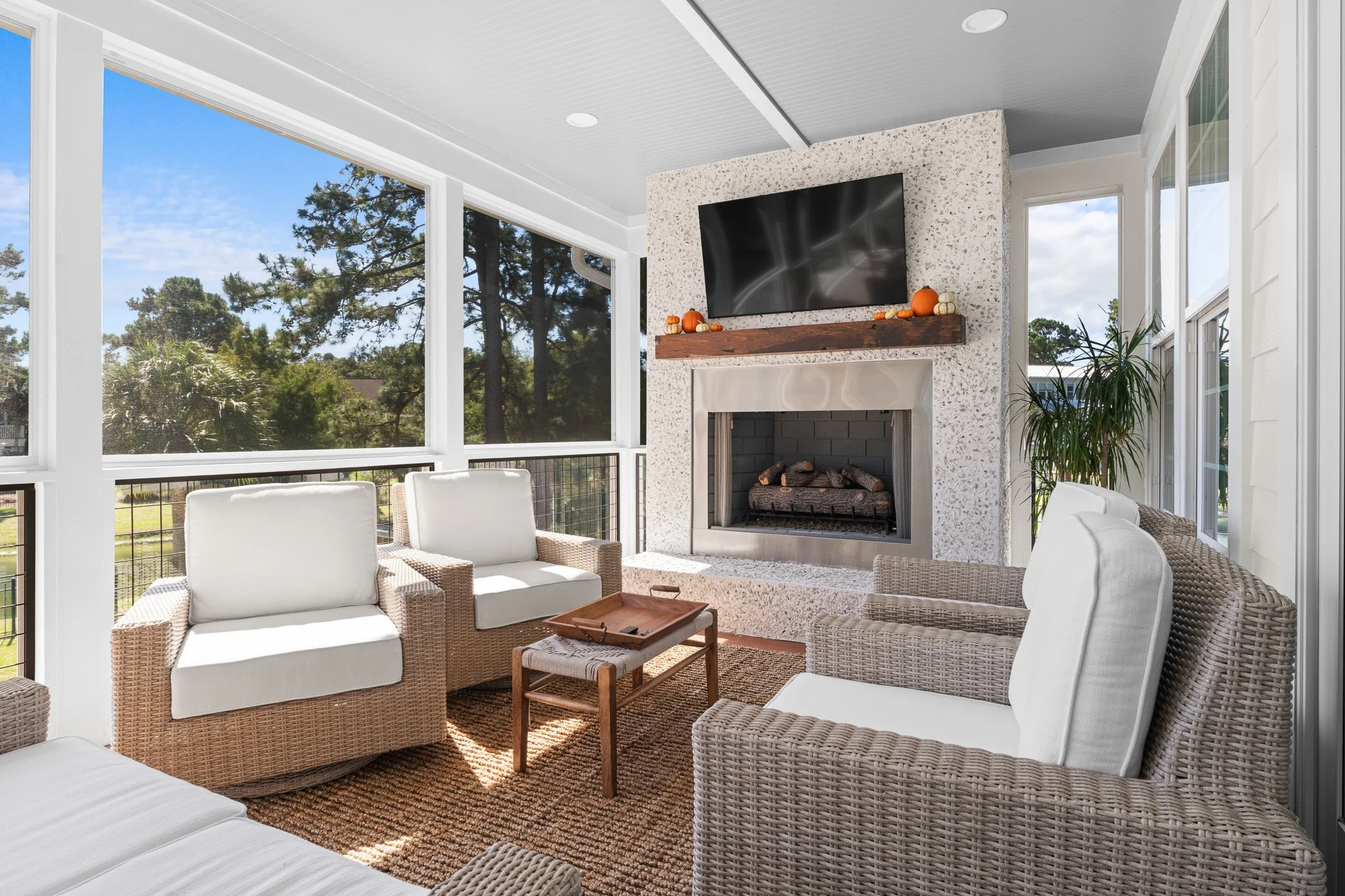 Sunroom with white wicker furniture around a rug, ceiling-high windows showing trees and blue sky, and a fireplace with a TV mounted above, decorated with small pumpkins.