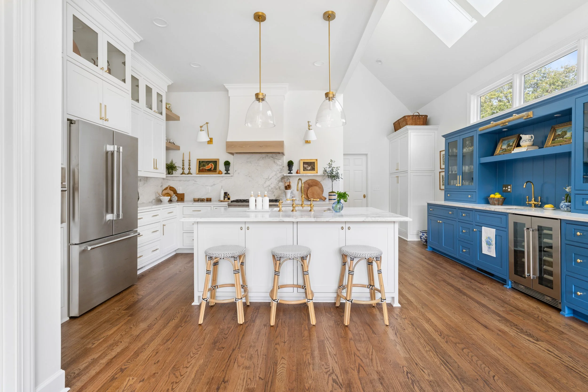 A bright kitchen with white cabinets and a central island with three barstools. A blue hutch with gold hardware stands against the wall, and stainless steel appliances, including a refrigerator, are visible. The kitchen features wooden flooring and s