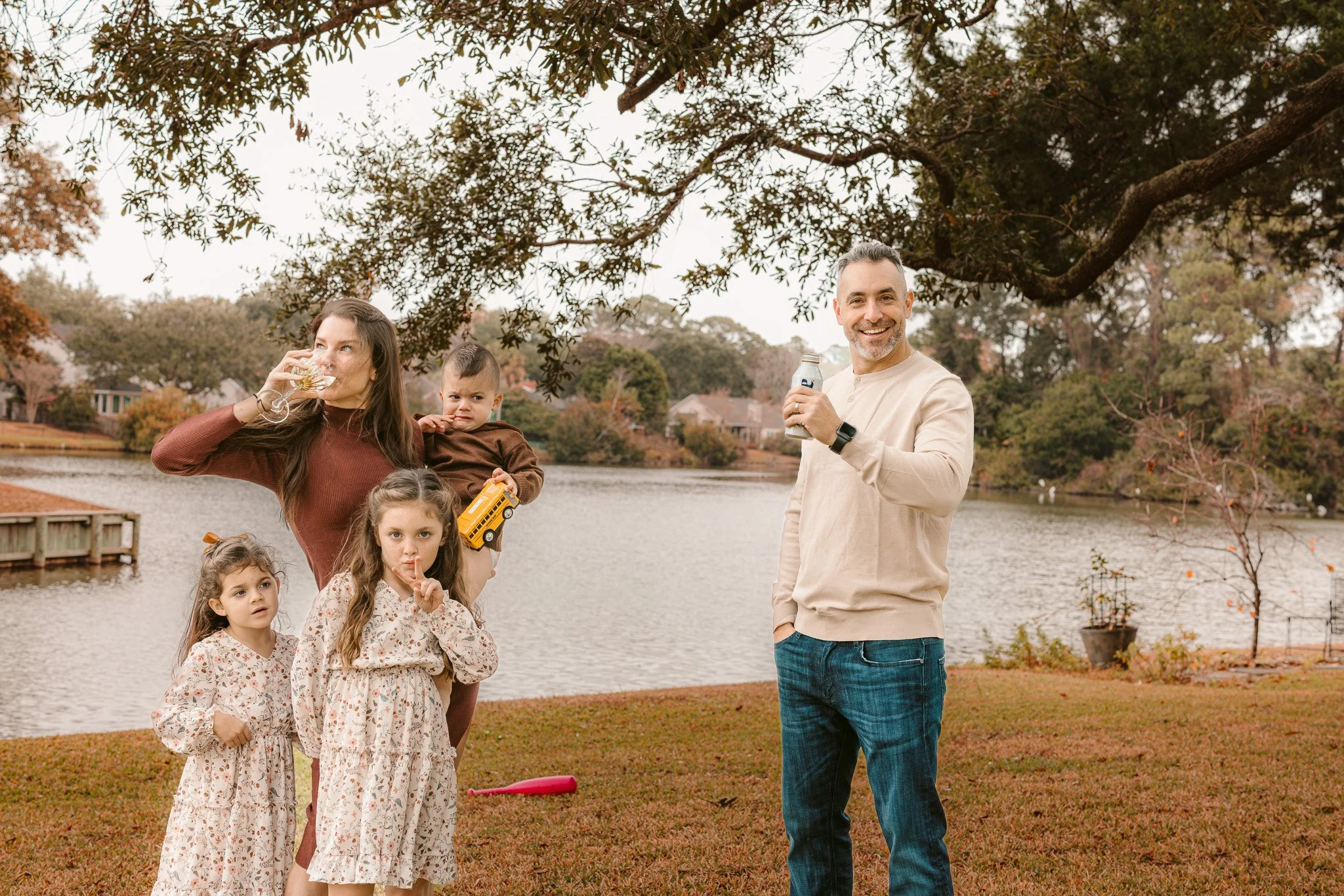 Family of five enjoying time outdoors by a lake in autumn. Two young girls in floral dresses, woman with a kid, and man smiling with a drink.