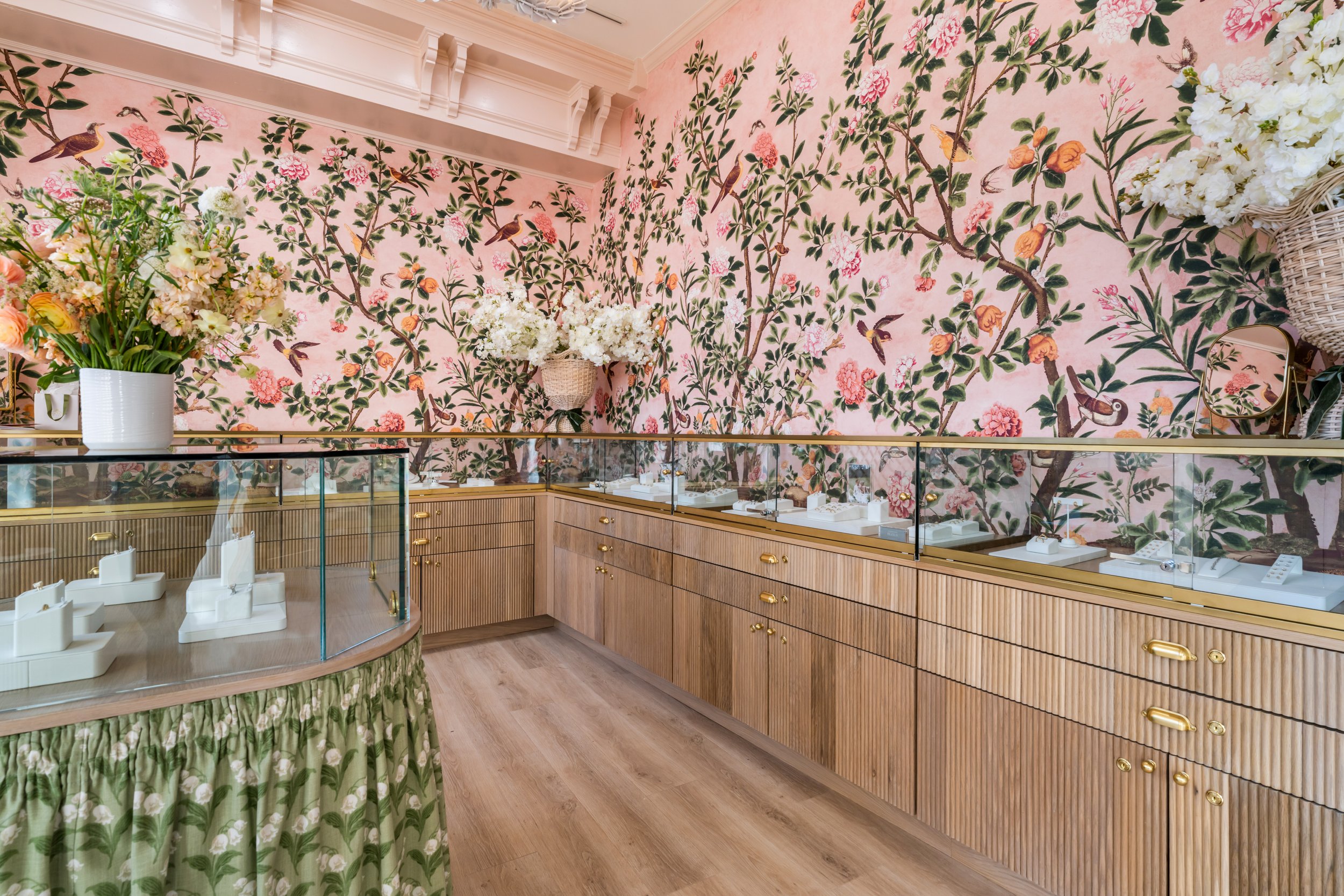Interior of a jewelry store with a pink floral wallpaper featuring birds, glass display cases with jewelry, wooden cabinets, large flower arrangements, a small mirror, and a green floral-printed curtain.
