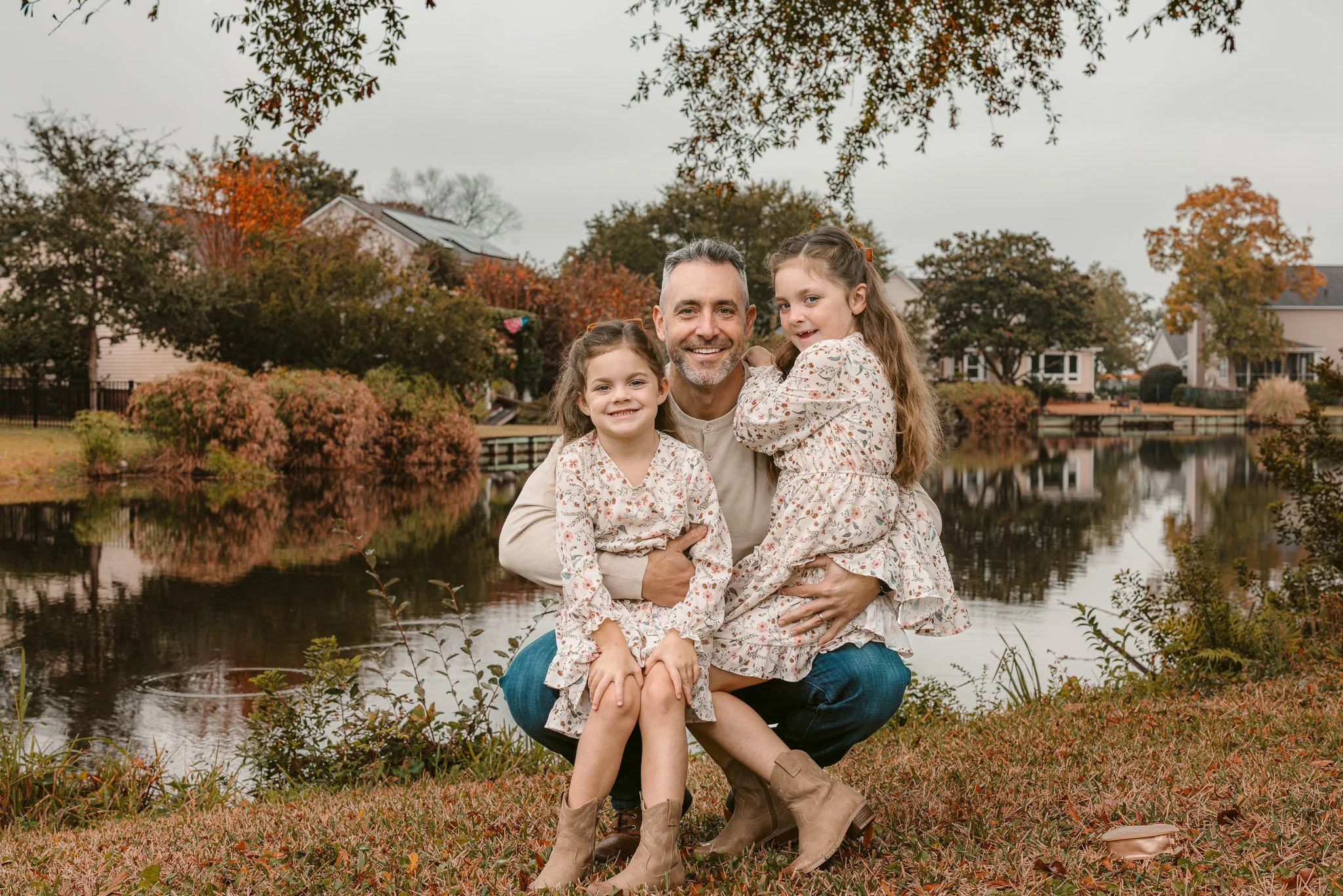 A man with two young girls on a grassy area by a lake during autumn. All three are smiling and wearing matching floral dresses. Trees with fall foliage and houses are in the background.