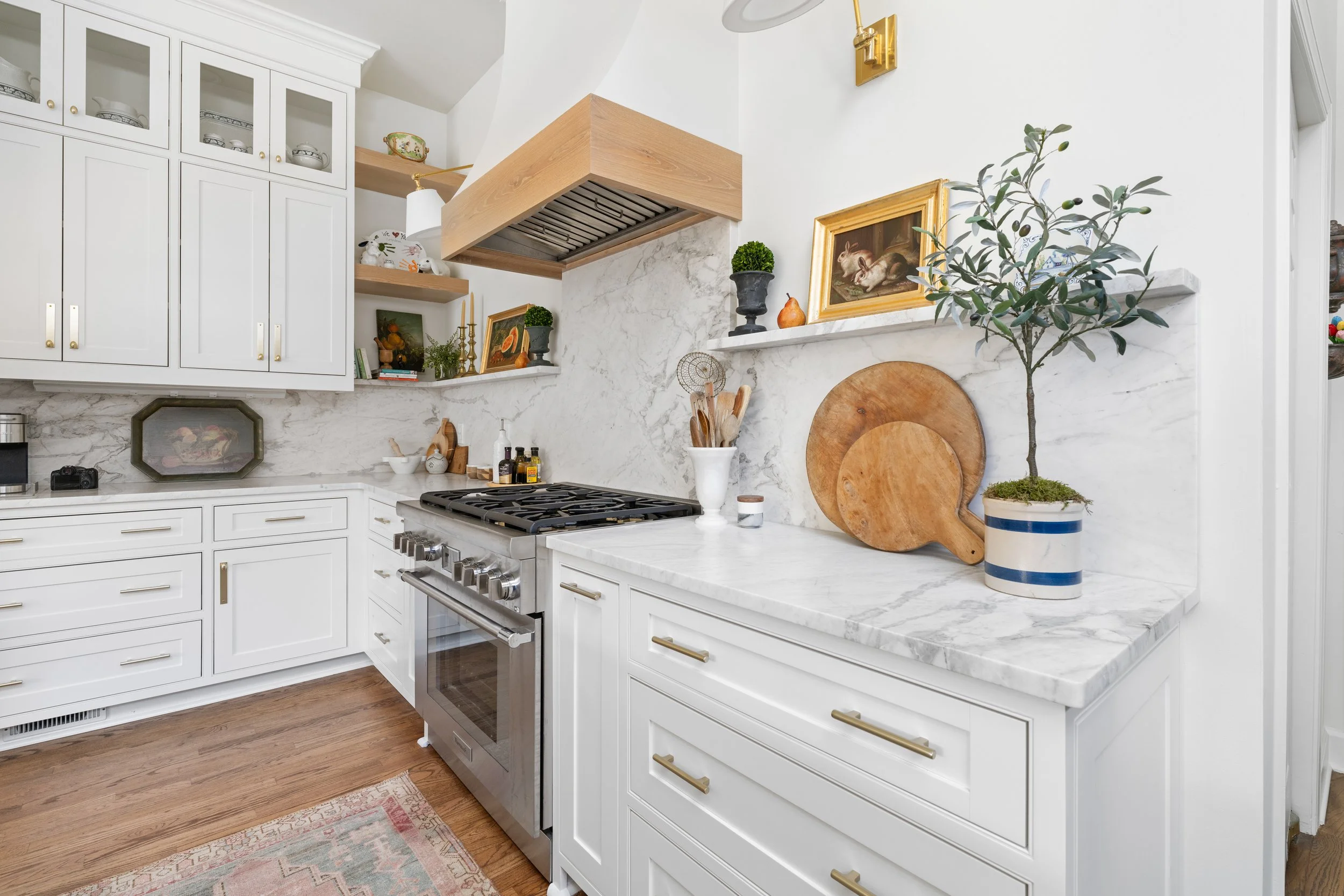 A kitchen with white cabinets, a stainless steel stove, and marble countertops. Decor includes framed art, candles, small plants, wooden cutting boards, and a potted olive tree. The space is bright with white walls and gold accents.