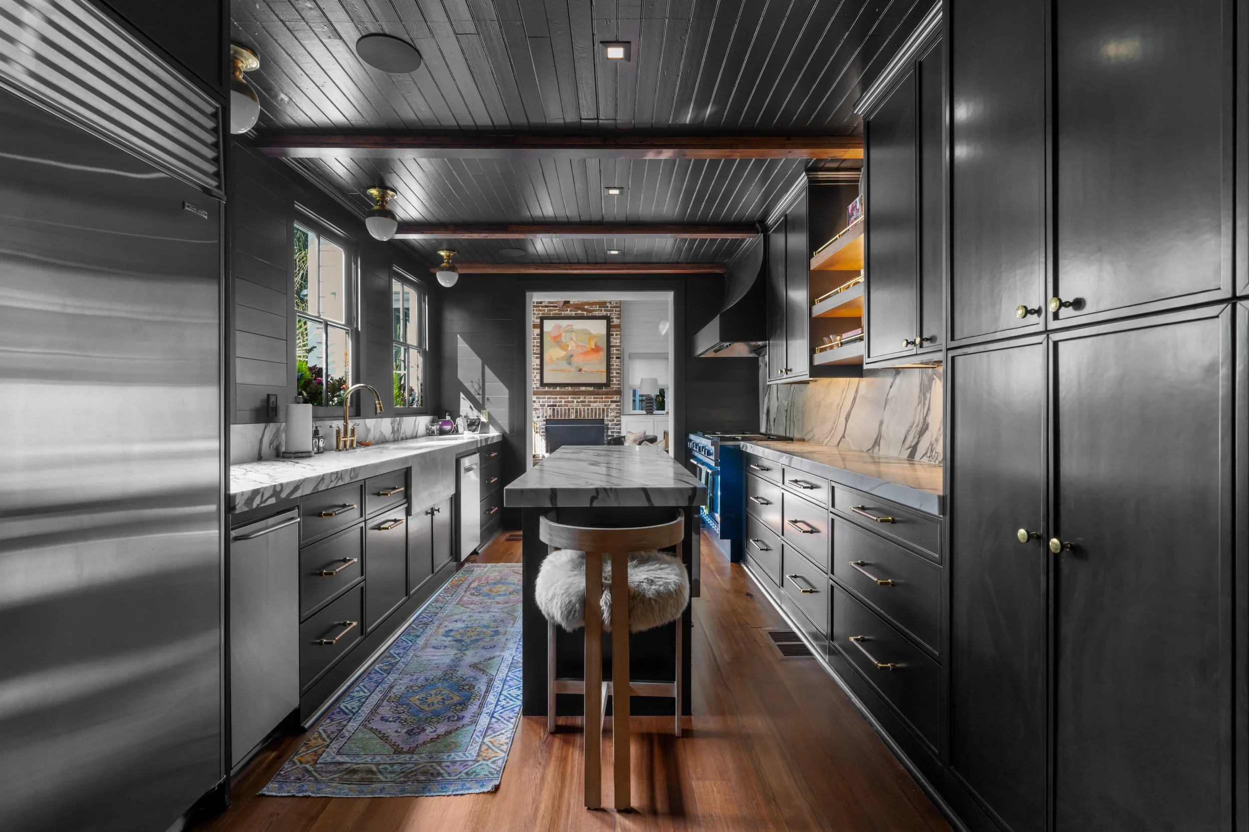 Modern black kitchen with white marble countertops, wooden floor, and a central island with a furry stool. Features large windows, ceiling lights, and open shelving with dishes, with a fireplace and artwork visible in the background.