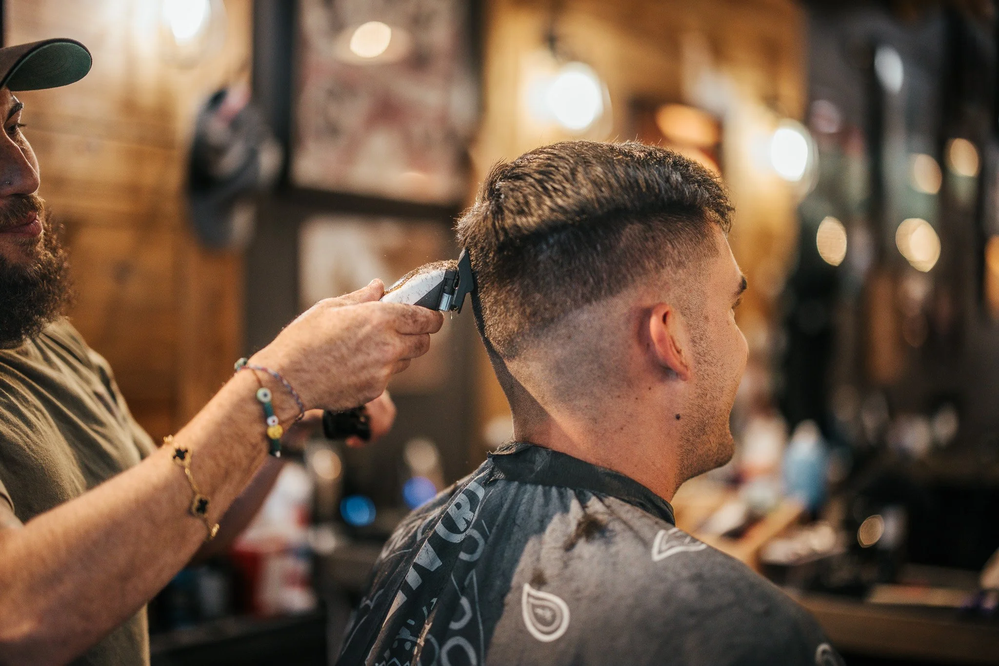 A barber trims a young man's hair with electric clippers inside a barbershop with wooden walls and various decorations.