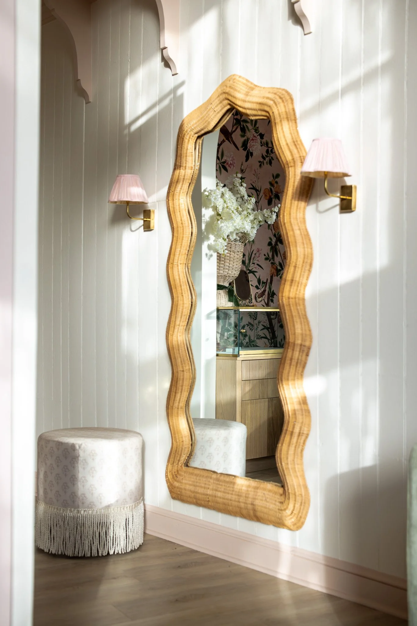 A tall, wavy-edged mirror with a wooden frame hanging on a light-colored panel wall. On each side of the mirror, there are two wall-mounted pink lamps with gold fixtures. Below the left lamp, there is a round ottoman with a light floral pattern and a