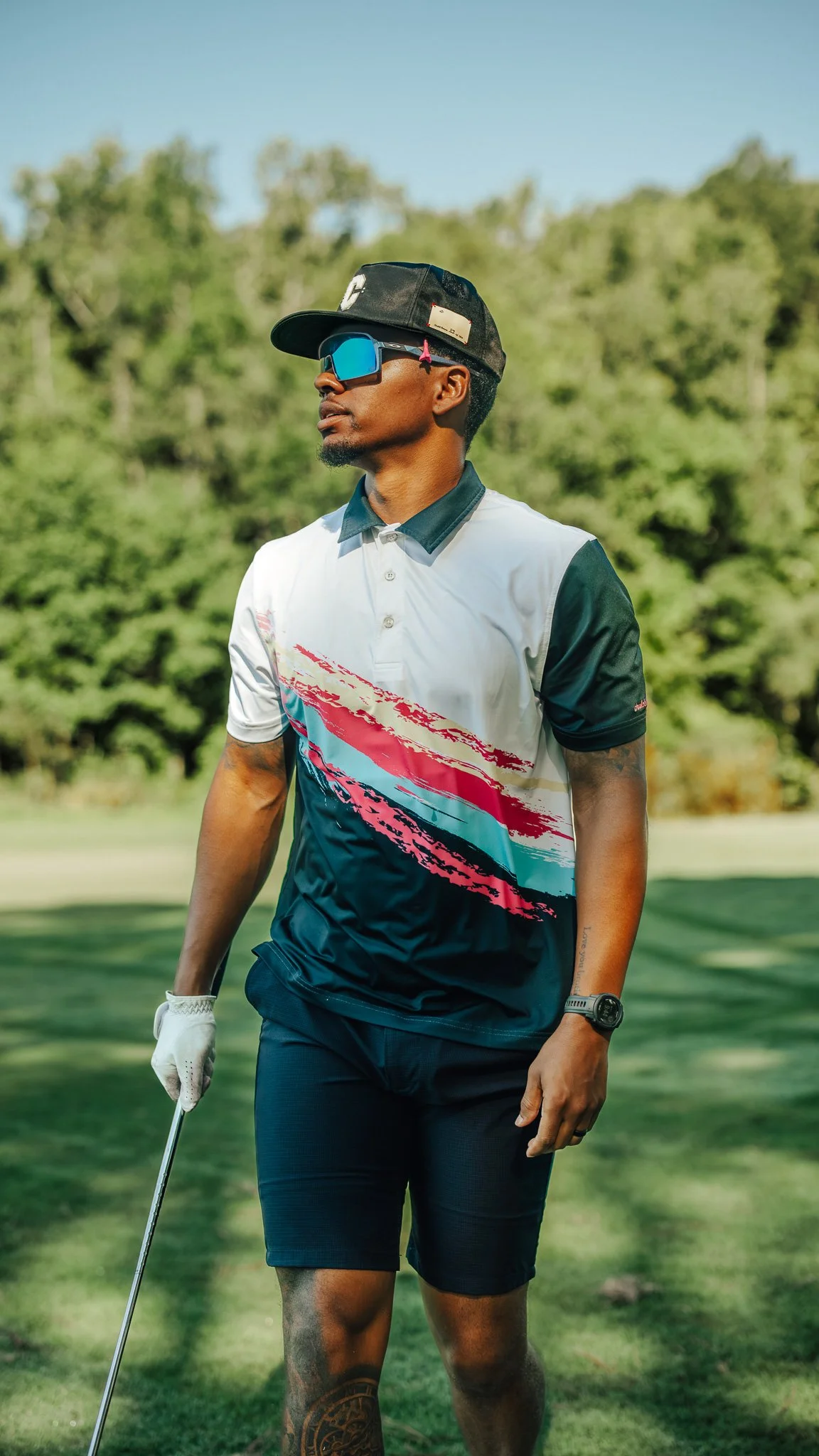 A man standing on a golf course holding a golf club, wearing sunglasses, a black cap, a white and colorful polo shirt, black shorts, and a watch.