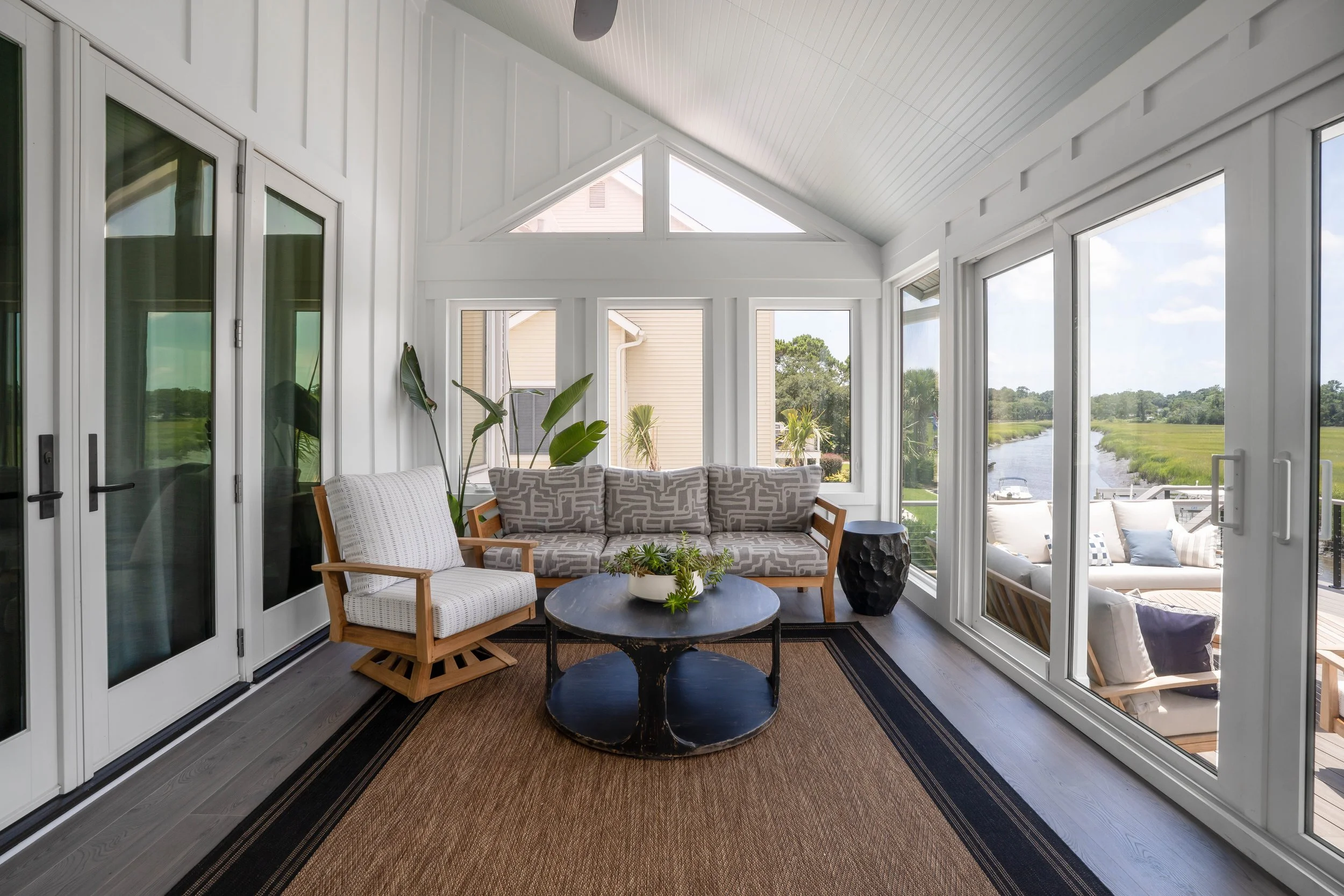 Bright sunroom with large windows overlooking a waterway, containing a patterned sofa, armchair, black coffee table with a plant, and potted plants in the background.